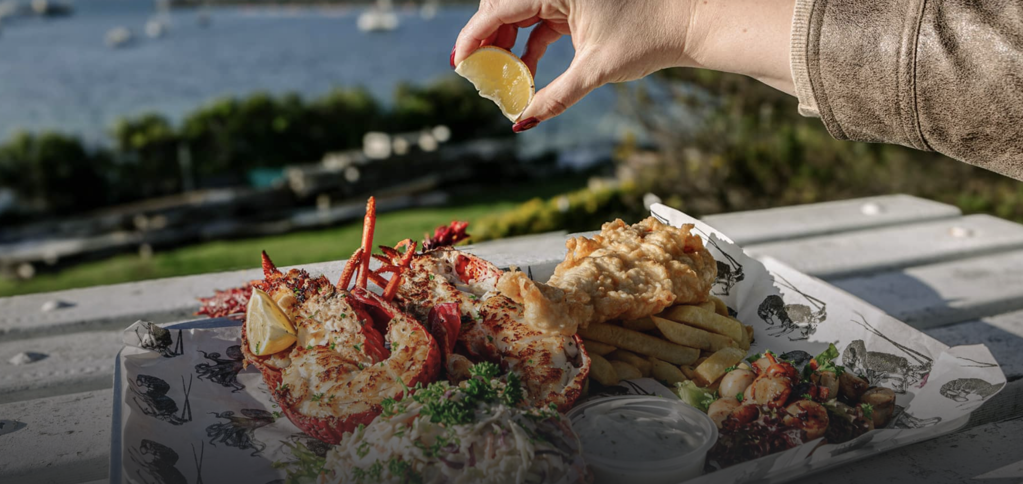 A seafood platter with lobster, fried fish, french fries, seafood salad, and a side of sauce on a white tray outdoors near a body of water, with a hand squeezing lemon over the lobster.