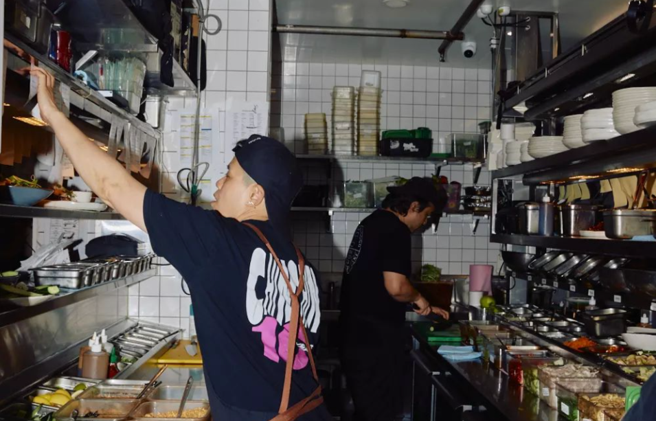 Two restaurant kitchen staff working at their stations with shelves of dishes and ingredients around them.