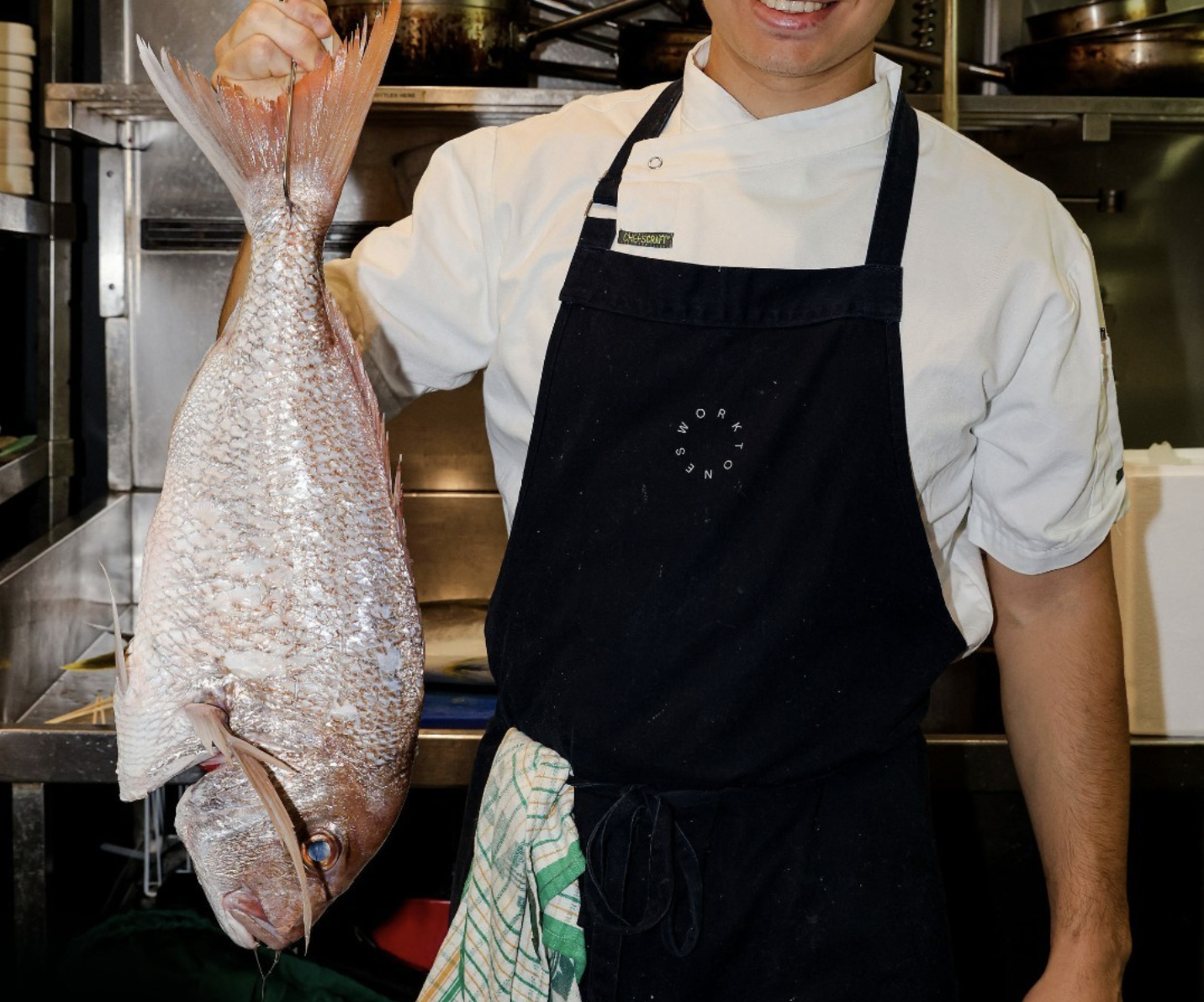 A person dressed as a chef in a white chef's coat and black apron, holding a large fish by its tail inside a commercial kitchen.