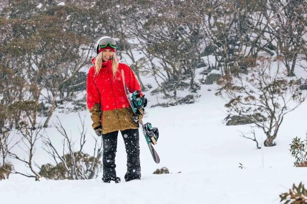 A woman standing in snow holding a snowboard, wearing a red and brown winter jacket, black pants, goggles, and a helmet, smiling during snowfall in a winter landscape with trees in the background.