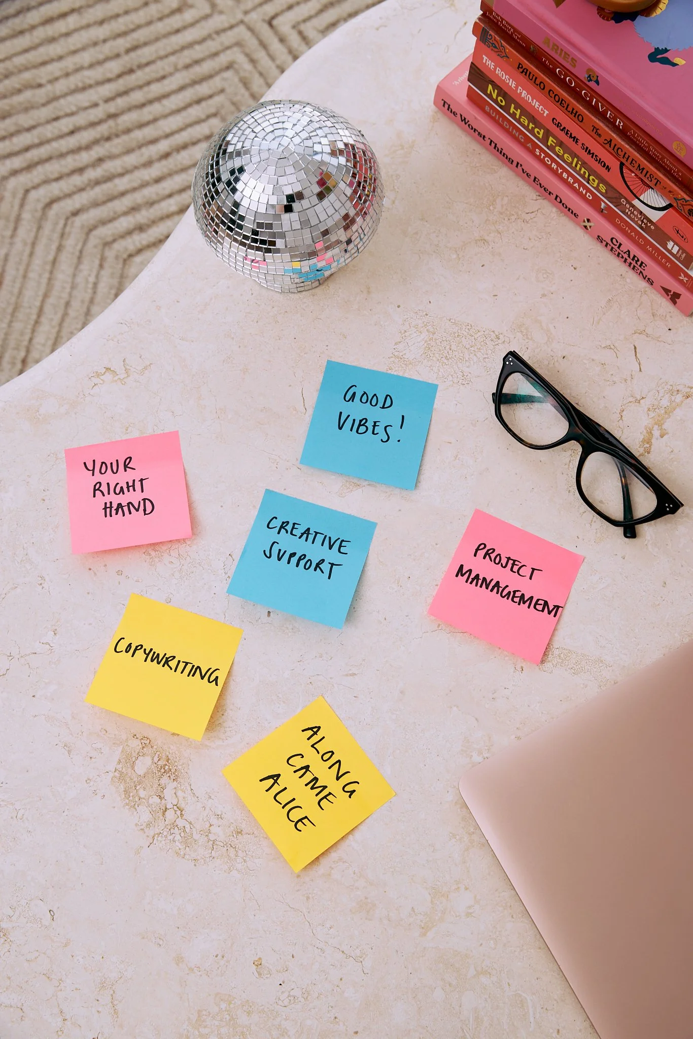 Colorful sticky notes with motivational phrases and tasks on a white desk, alongside a pair of black glasses, a stack of books, and a small disco ball.