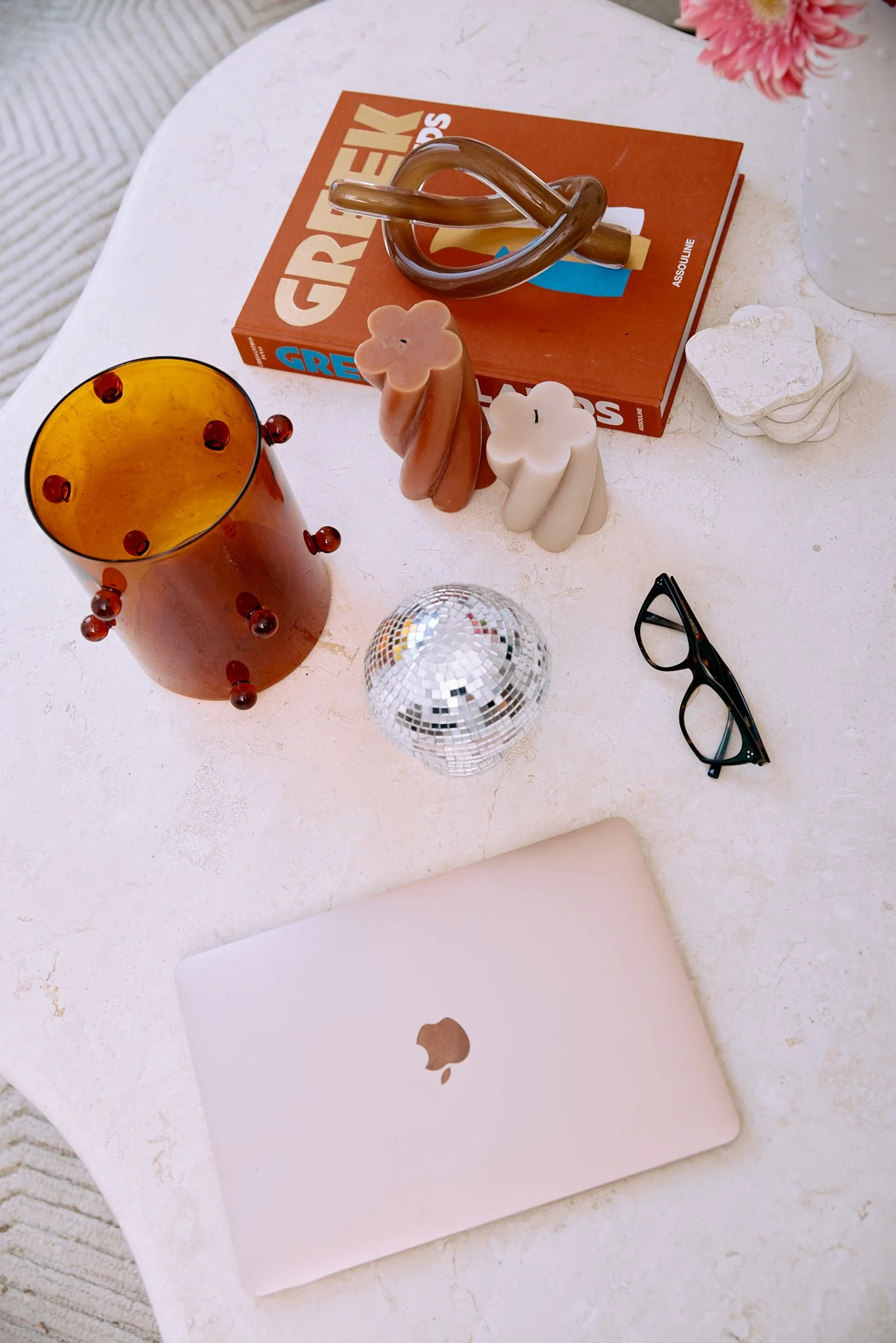 A white table with a closed pink MacBook, a small disco ball, black glasses, an orange glass with red beads, two textured candles shaped like people, a brown book titled 'GREAT GAY BOOKS', a clear glass sculpture, and a white vase with pink flowers.
