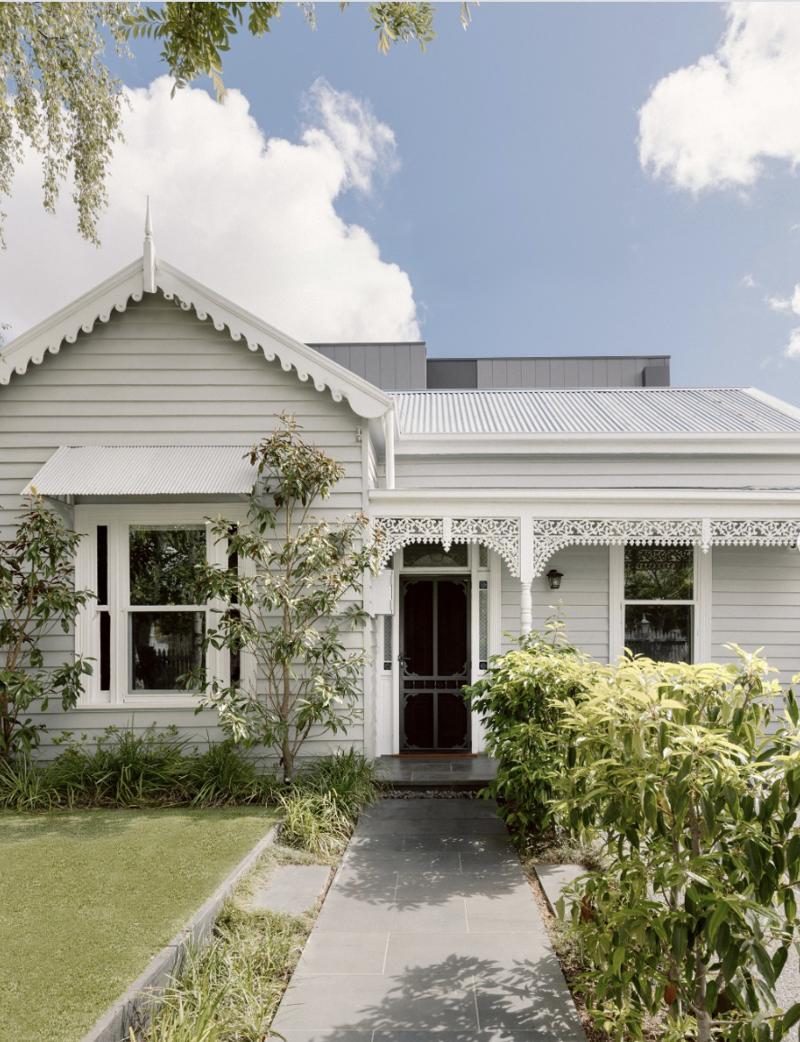 White house with Victorian architectural details, front walkway, green plants and trees, cloudy sky.