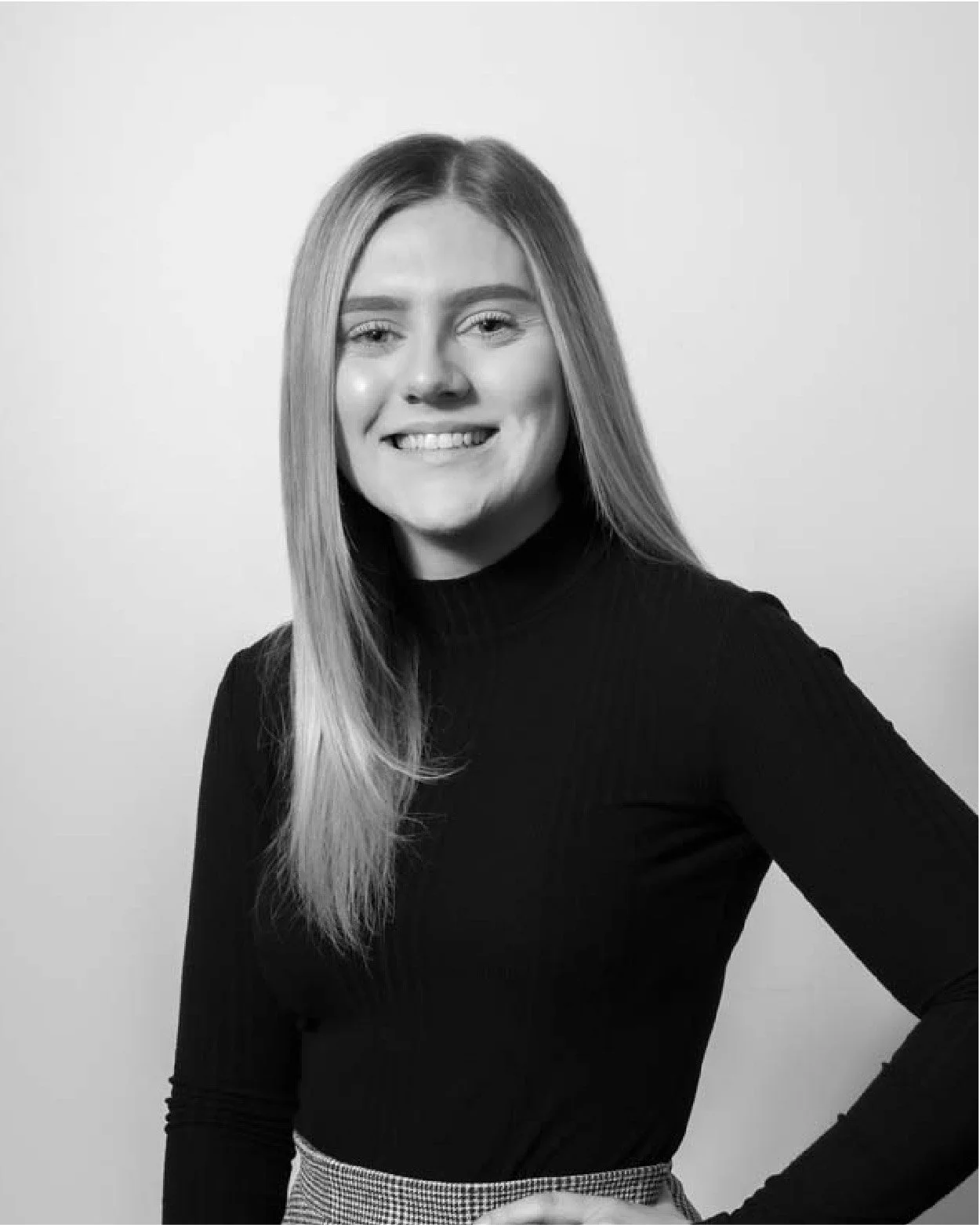 Black and white portrait of a smiling woman with long straight hair, wearing a dark turtleneck and checkered pants, standing against a plain background.