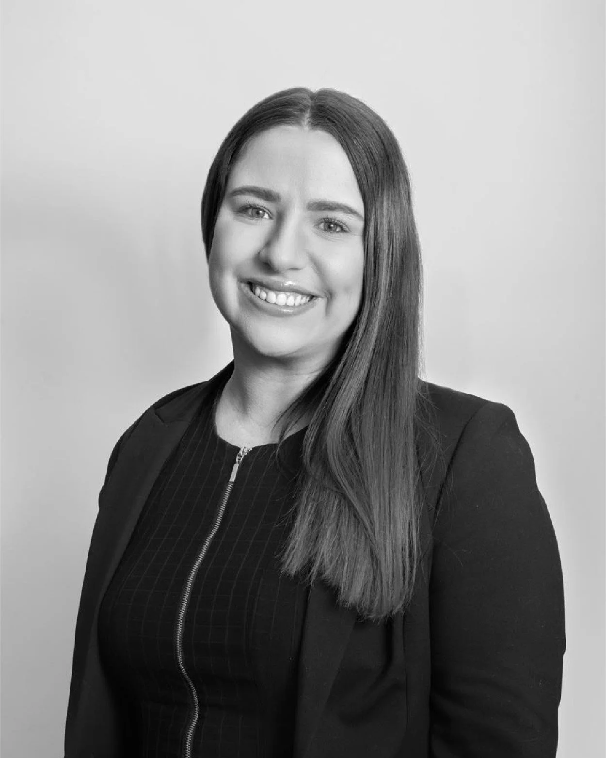 Black and white photo of a woman with long straight hair, smiling, wearing a dark blazer over a zipper top, standing against a plain background.