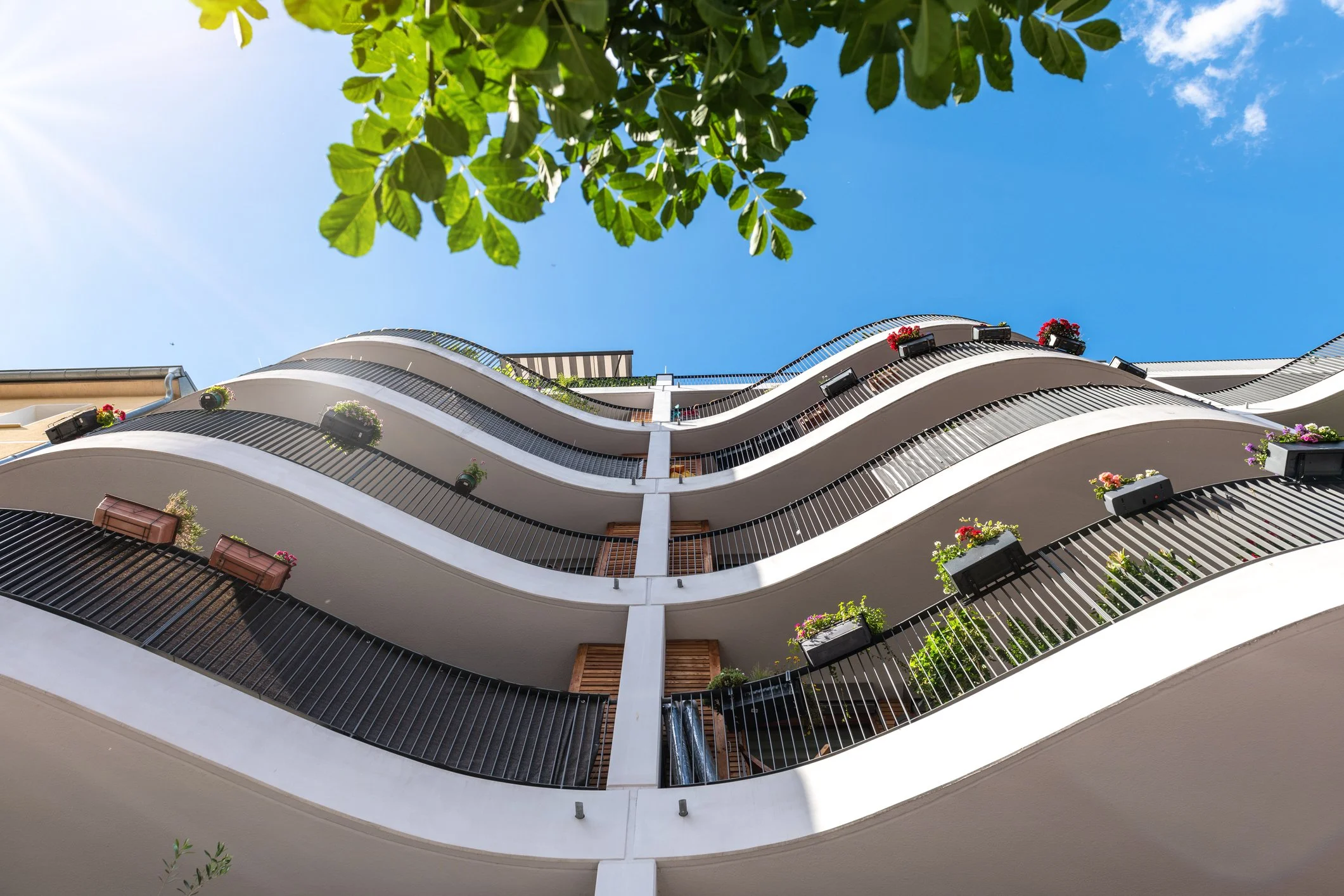 Low-angle view of a modern apartment building with wavy balconies, flower pots, and a blue sky with some clouds.