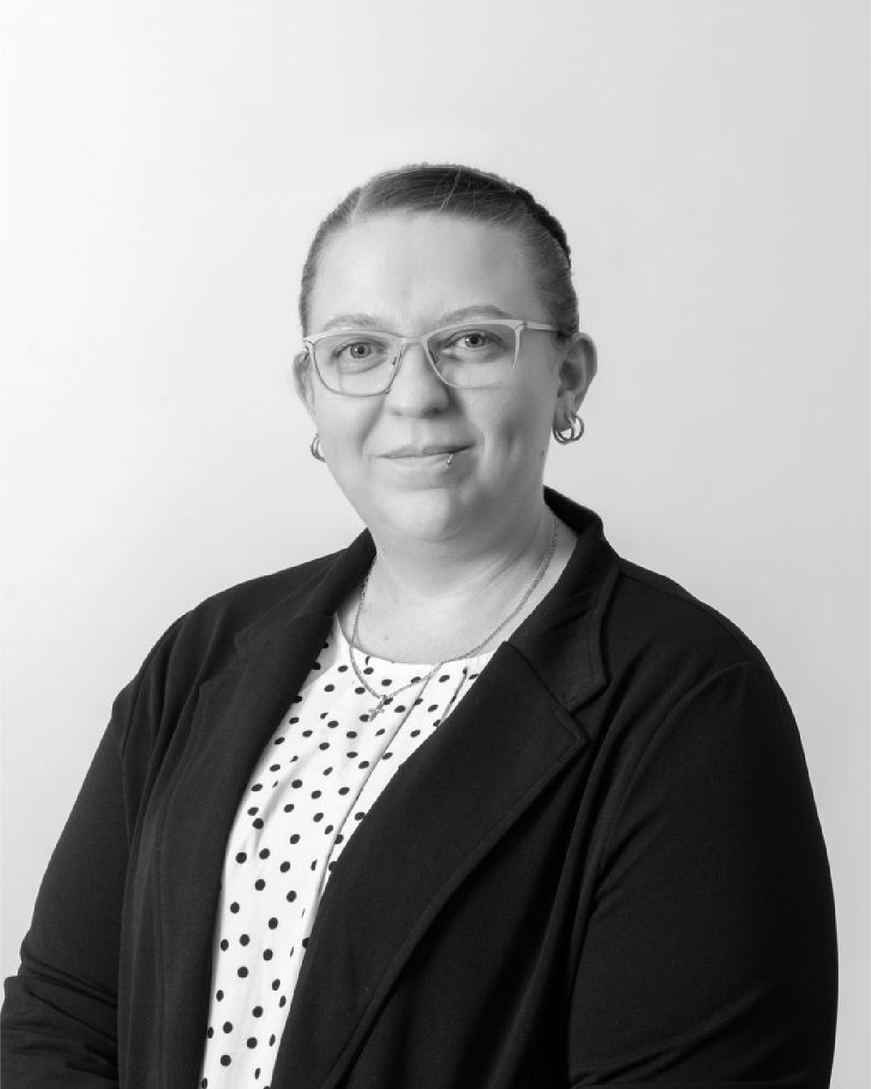 Black-and-white portrait of a woman wearing glasses, earrings, and a dark jacket, smiling slightly against a plain white background.