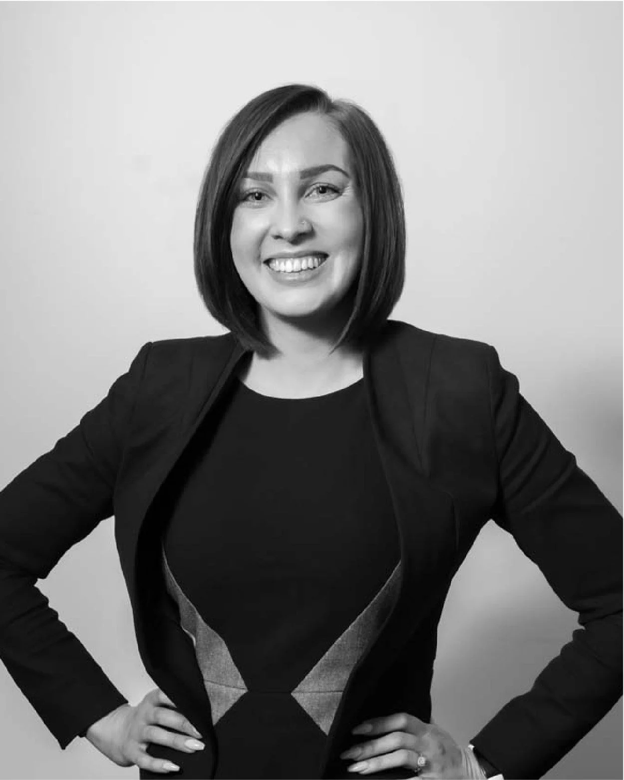 A woman with shoulder-length hair, smiling, wearing a dark blazer over a patterned top, with her hands on her hips, black and white portrait.