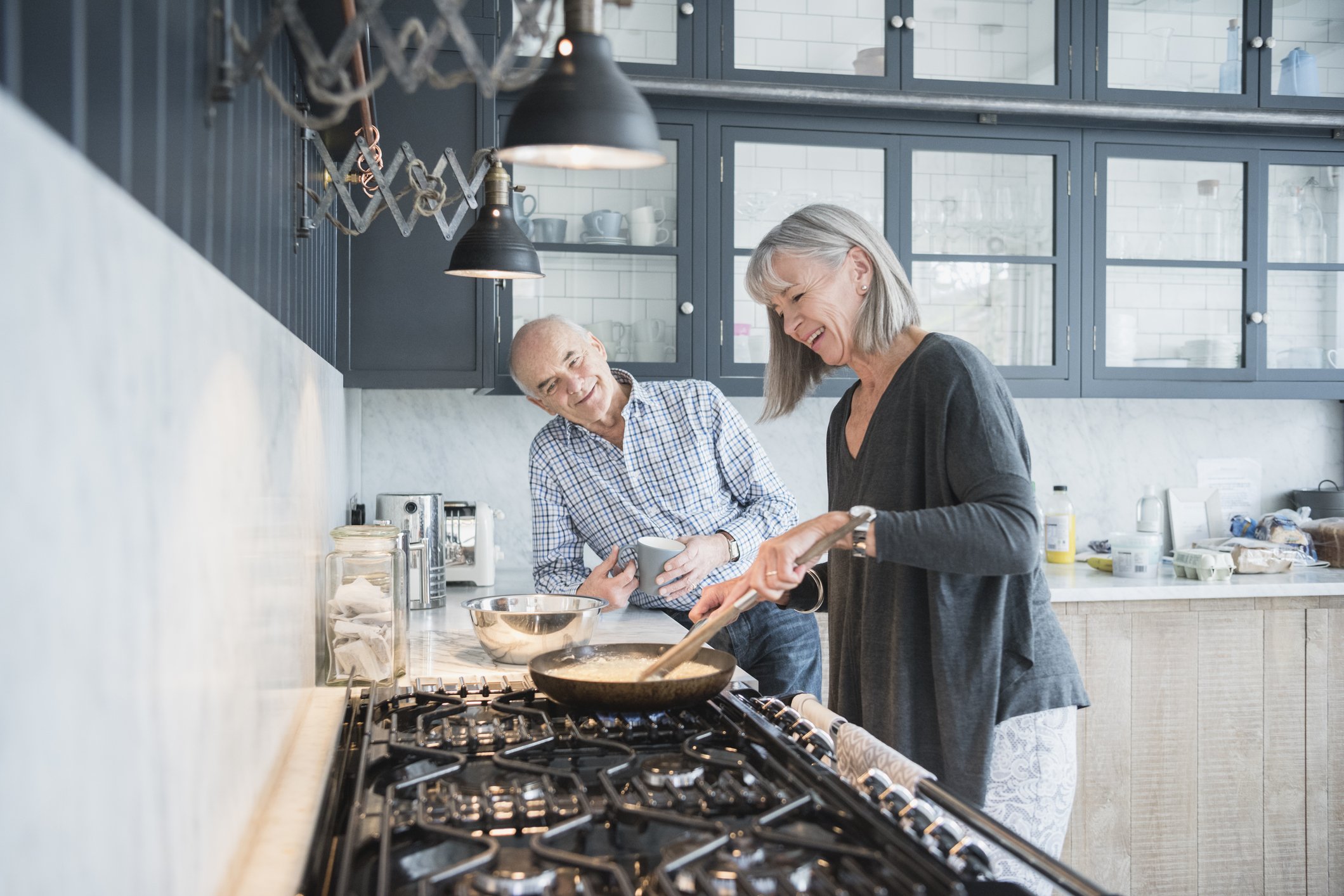 A smiling older man and woman cooking together in a modern kitchen, with the woman stirring a frying pan and the man holding a mug, both appearing happy and engaged.