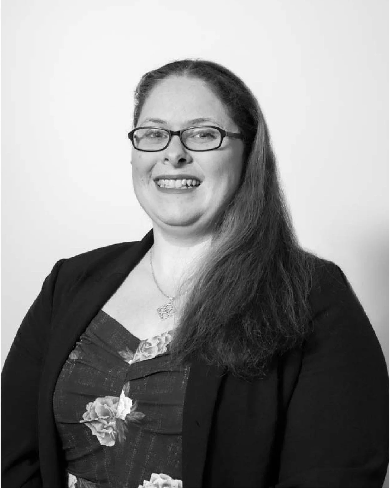 Black and white portrait of a woman with glasses, long hair, wearing a floral top and a blazer, smiling at the camera.