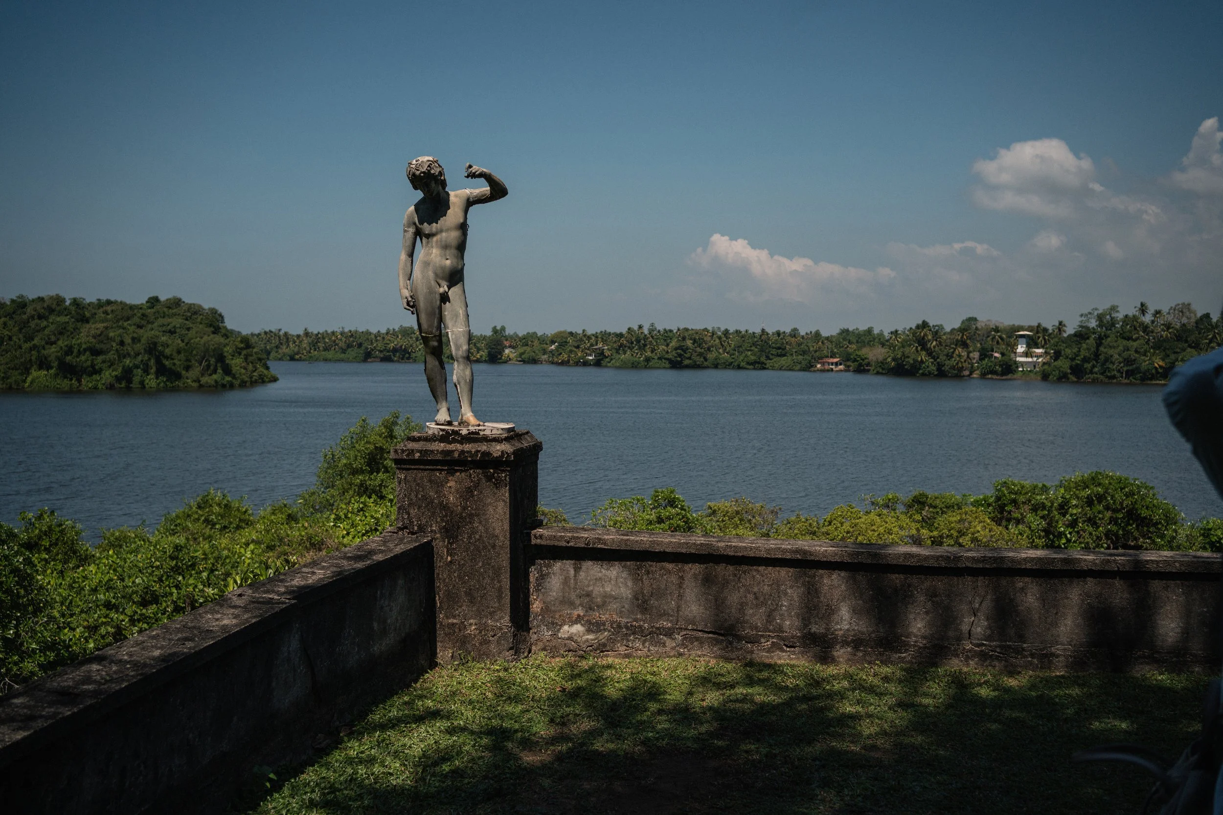 Statua di un uomo nudo con un braccio alzato, situata su un parapetto di pietra, con vista su un lago e una natura verdeggiante sotto un cielo blu con alcune nuvole.