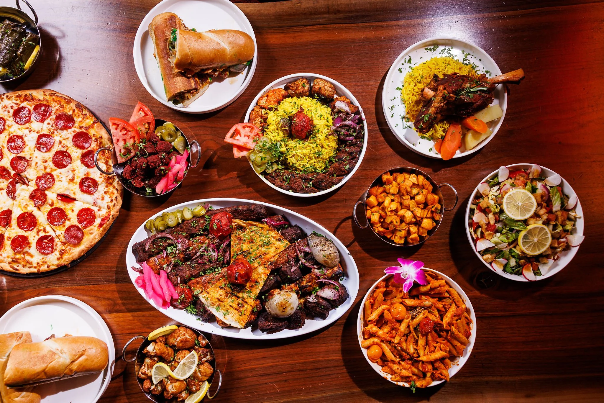 A variety of Indian and Western dishes on a wooden table, including pizza, naan, rice dishes, salads, grilled meats, and vegetable curries.