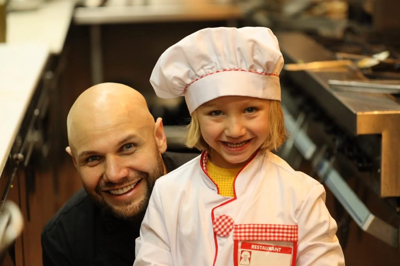 A young girl dressed as a chef, smiling, with a man in a restaurant kitchen. The girl is wearing a white chef hat and coat with red checkered accents, and the man is bald with a beard, wearing black.