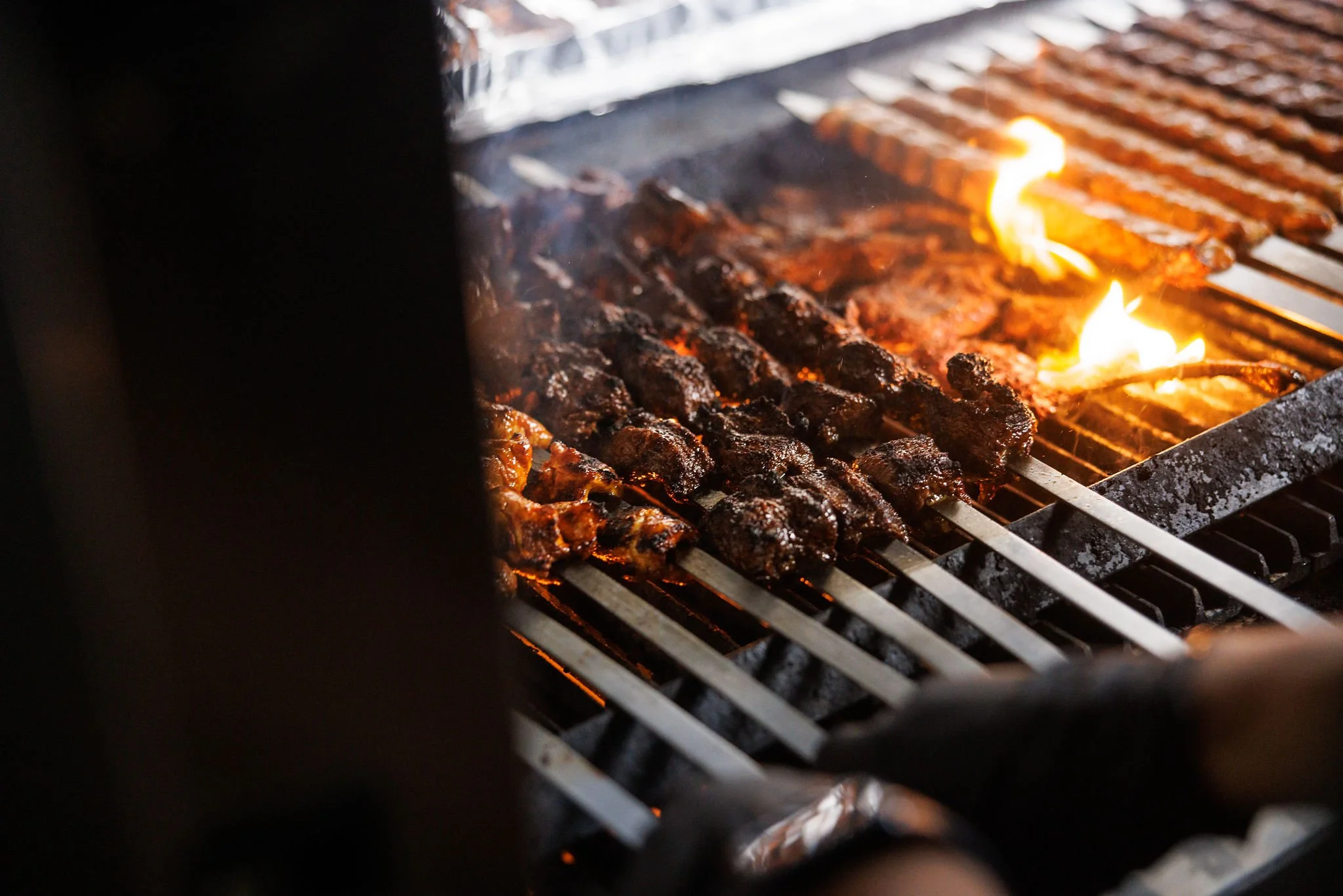 Skewers of meat being grilled on an outdoor barbecue with visible flames.