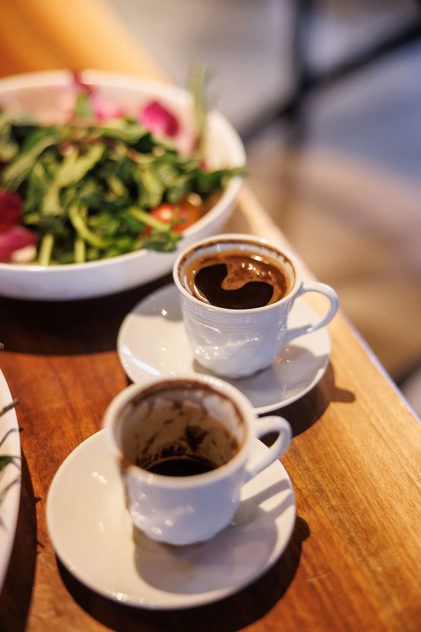 Two small white coffee cups filled with black coffee on a wooden table, with a bowl of salad in the background.