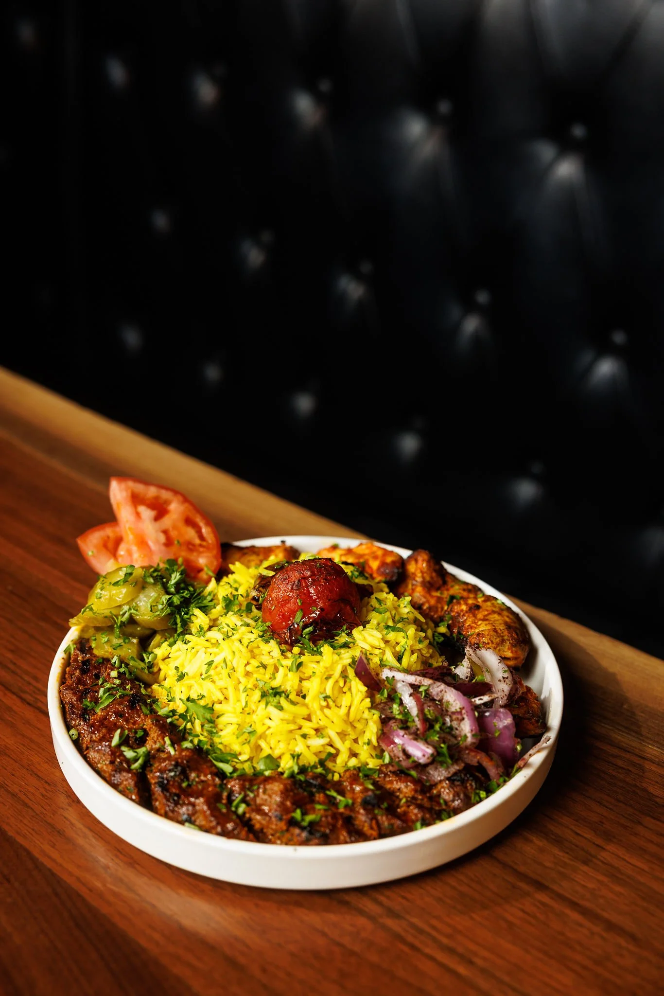 A bowl of Indian-style biryani rice served with grilled chicken, sliced onions, chopped tomatoes, and chutney, garnished with herbs, on a wooden table.