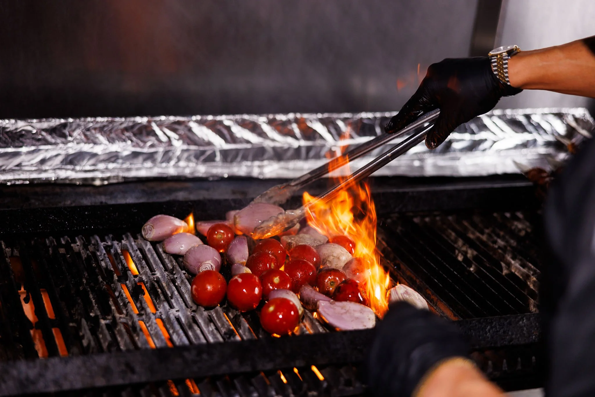 Person wearing black gloves and a silver watch using tongs to cook cherry tomatoes and onion slices on a grill with flames.