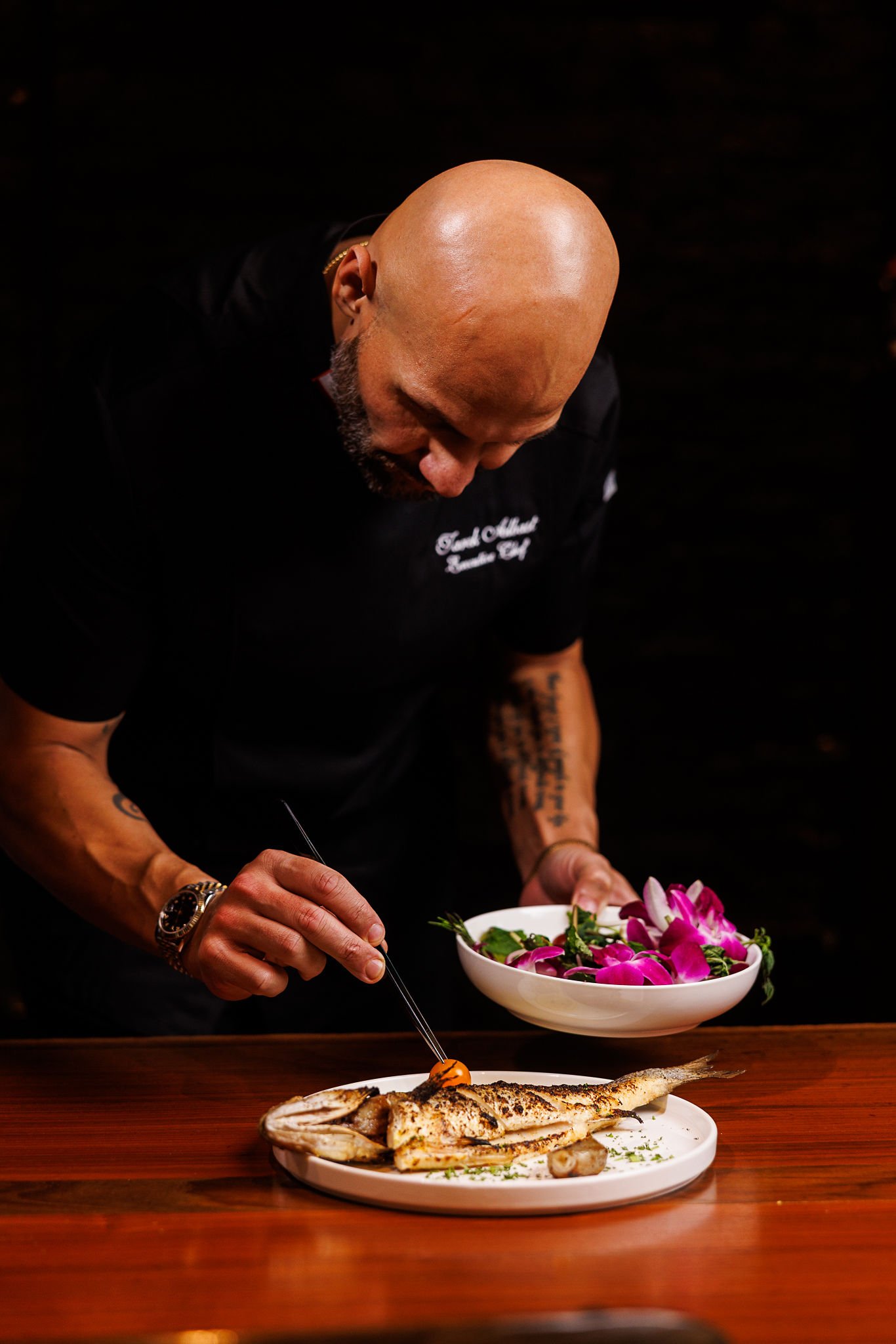 A man chef in black uniform garnishing a grilled fish on a white plate with herbs, holding a bowl of pink and purple flowers, standing at a wooden table