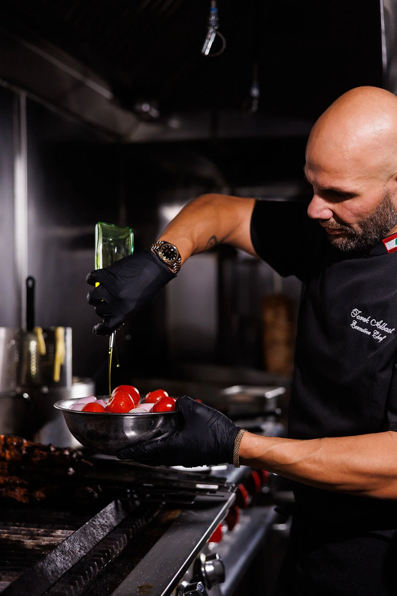 A chef with a bald head and beard wearing black gloves, a black shirt with embroidery, and a watch, is holding a black bowl of red tomatoes and pouring olive oil over them in a professional kitchen.