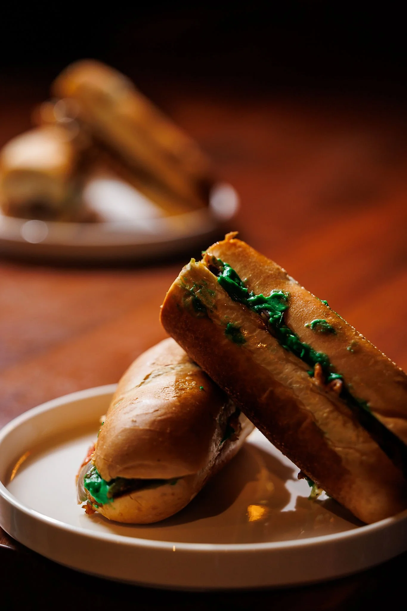 Close-up of two hot dogs with green relish on a white plate, with a blurred plate of more hot dogs in the background.