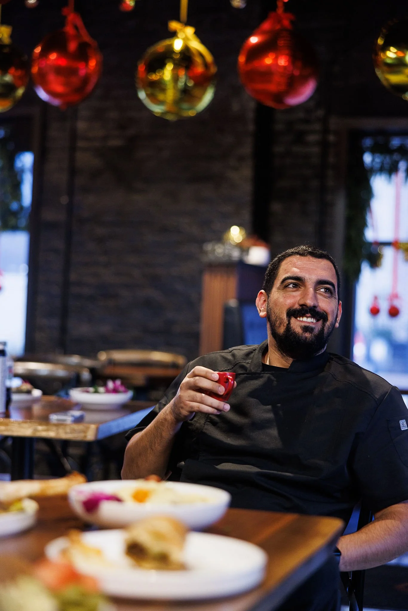 A man with dark hair and a beard, wearing a black chef's coat, smiling and holding a small red object, sitting at a table in a restaurant decorated with red and gold Christmas ornaments hanging from the ceiling.