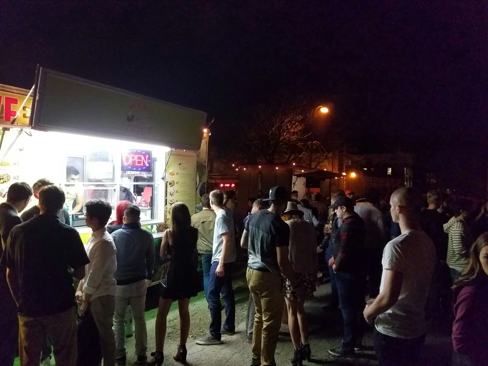 Crowd of people waiting in line at a food truck stand during nighttime, with some illuminated signs and streetlights in the background.