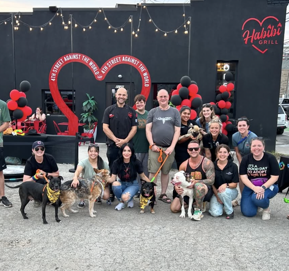 A group of people, including dogs and a cat, gathered outside a black building with red and black balloons and string lights. The building is called "Habibi Grill" with a heart-shaped logo. The group appears to be celebrating or participating in a community event.