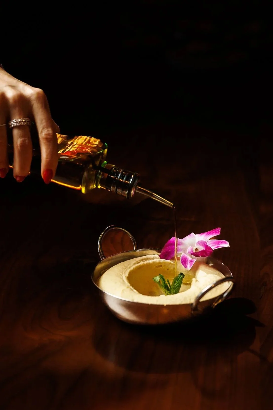 A hand pouring syrup or oil from a glass bottle onto a bowl of hummus garnished with pink flowers and herbs, on a dark wooden surface.