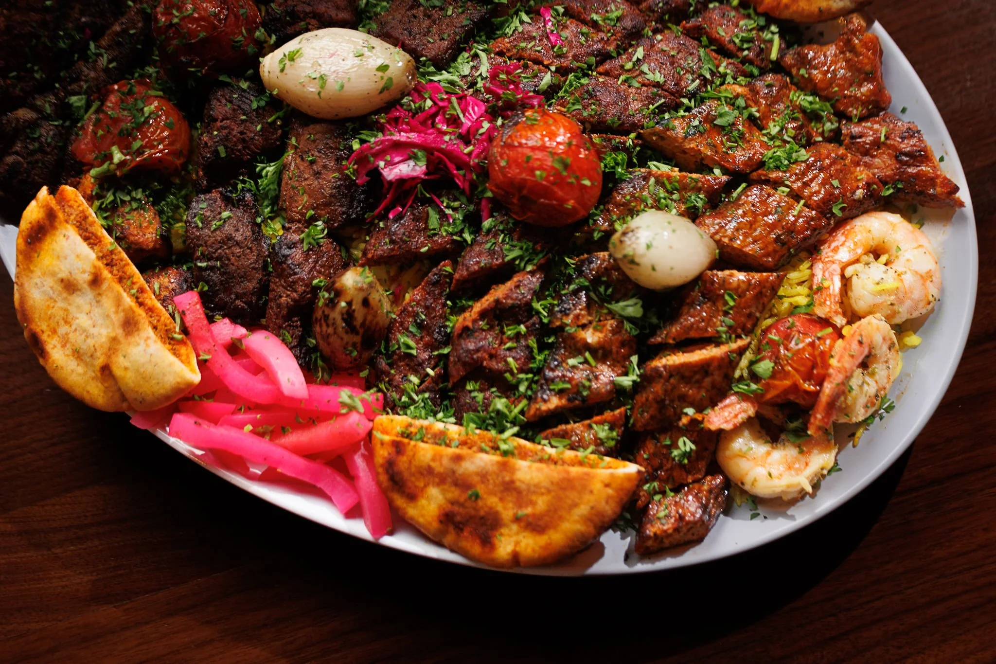 A large platter of assorted Mexican grilled meats including beef, shrimp, and sausage, garnished with herbs, pickled pink onions, and accompanied by grilled flatbread.