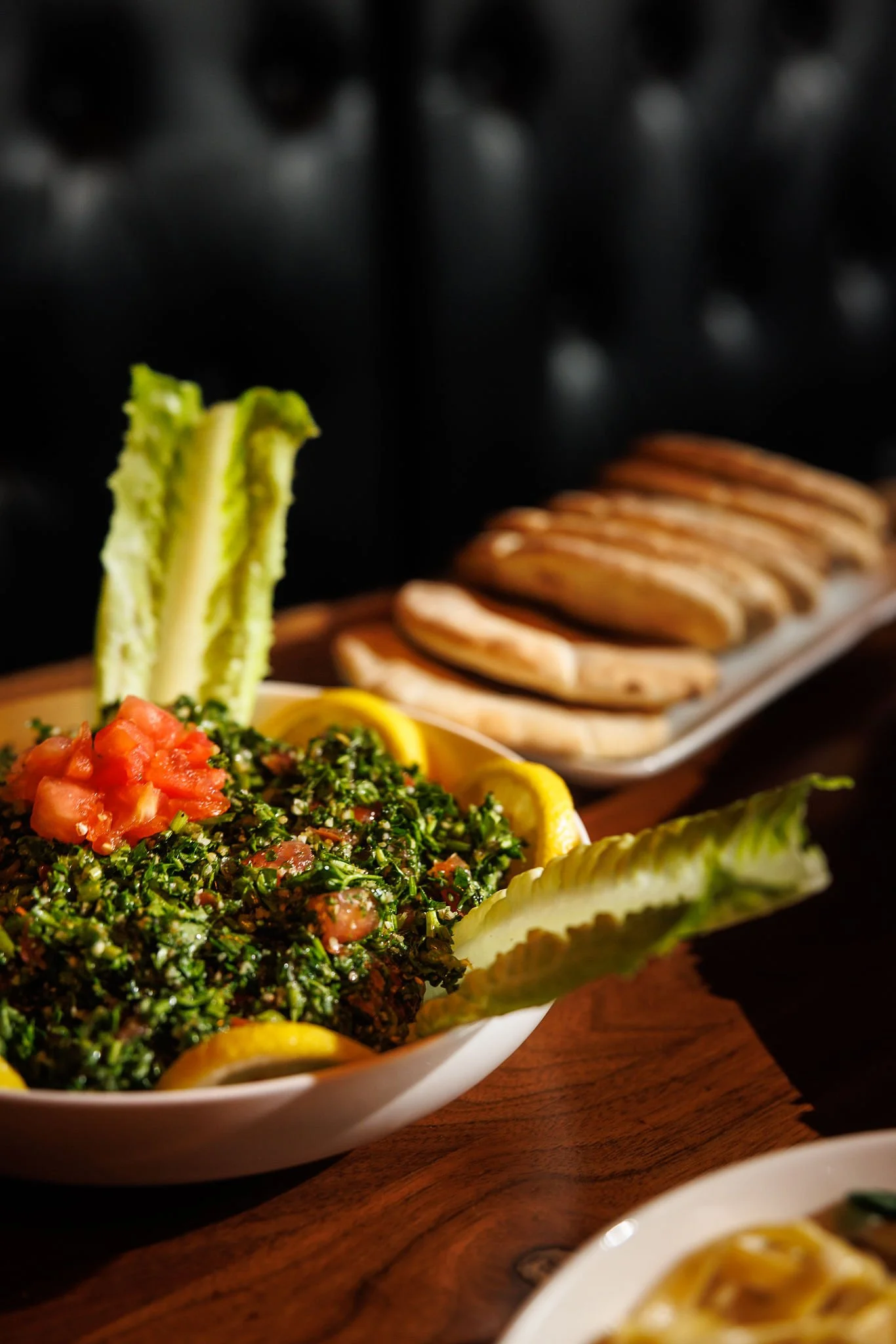 A platter of tabbouleh salad garnished with tomato and lemon slices, with celery sticks, and flatbread in the background.