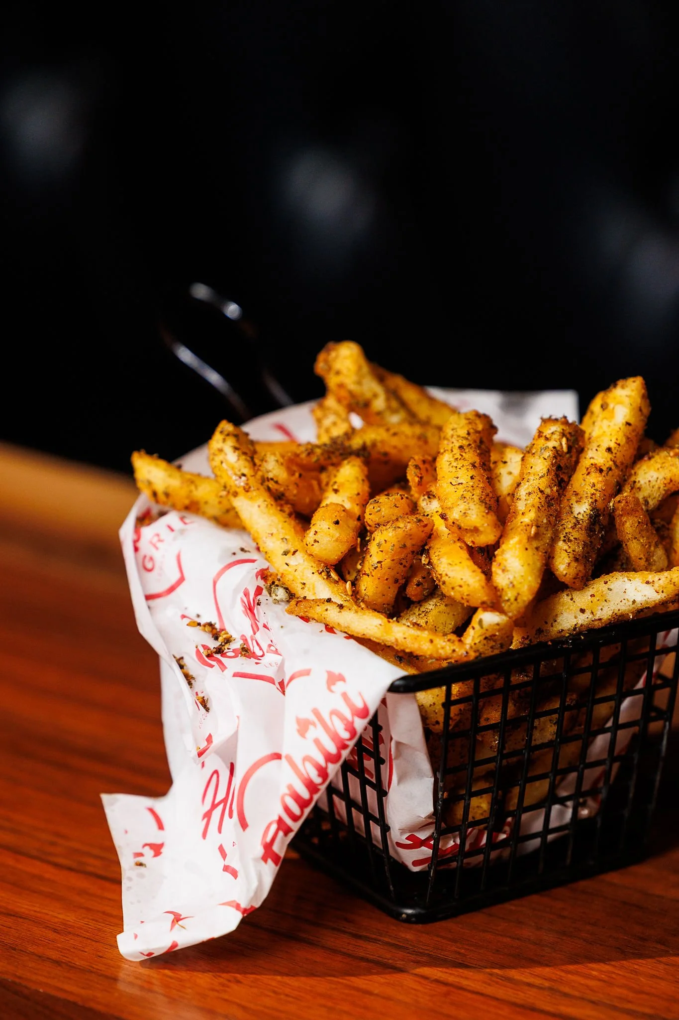 Basket of seasoned French fries on a wooden table, lined with branded paper.
