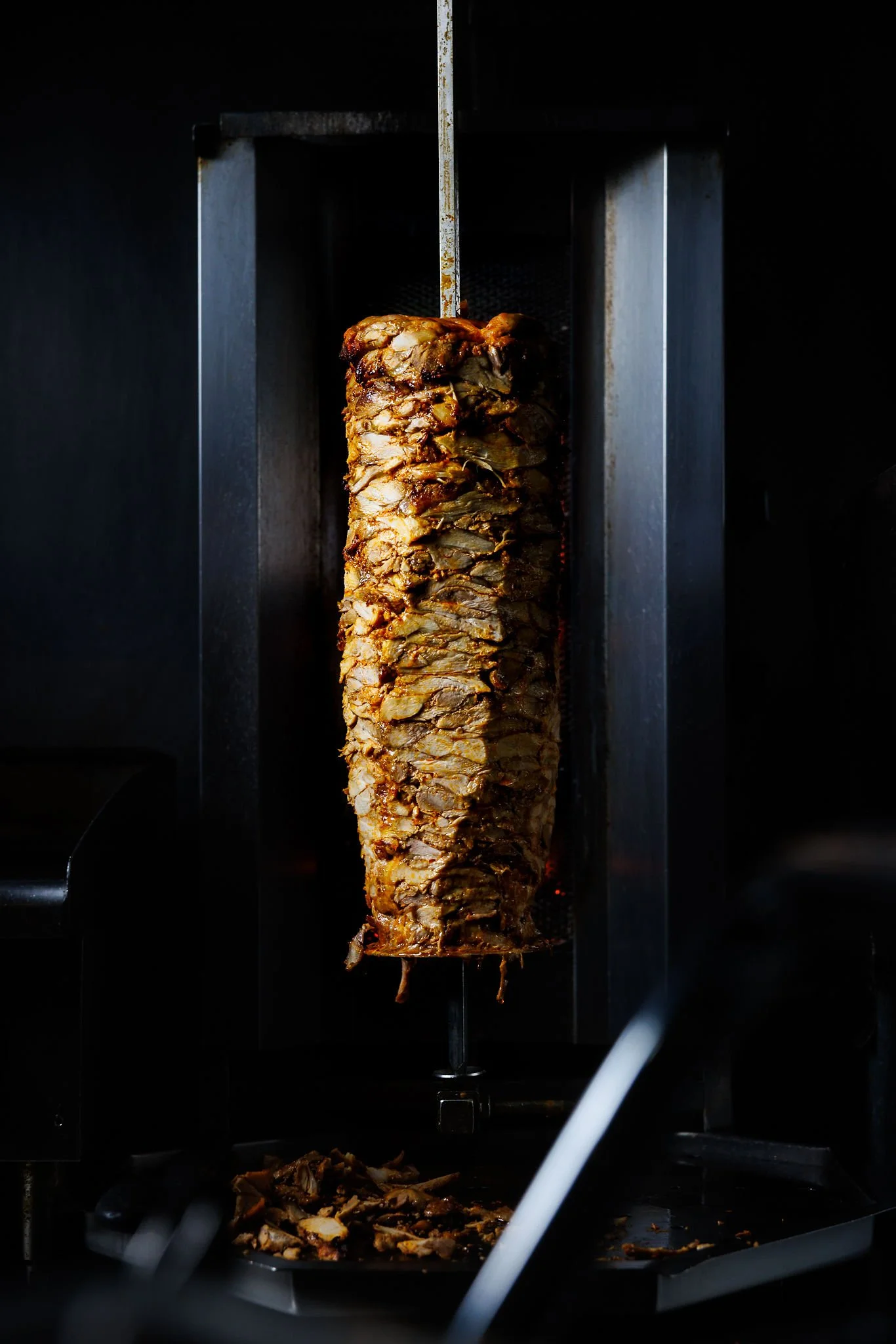 A vertical rotisserie meat, likely shawarma or gyros, cooking on a spit. The meat is sliced at the bottom, with some slices visible on a tray below.