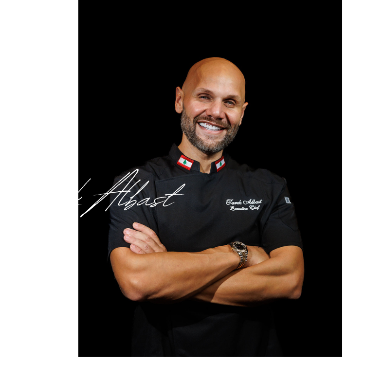 A smiling man with a beard and a bald head, wearing a black chef's coat with Lebanese flags on the collar, crossing his arms against a black background, with the name 'J' Albahst' written in white cursive text.