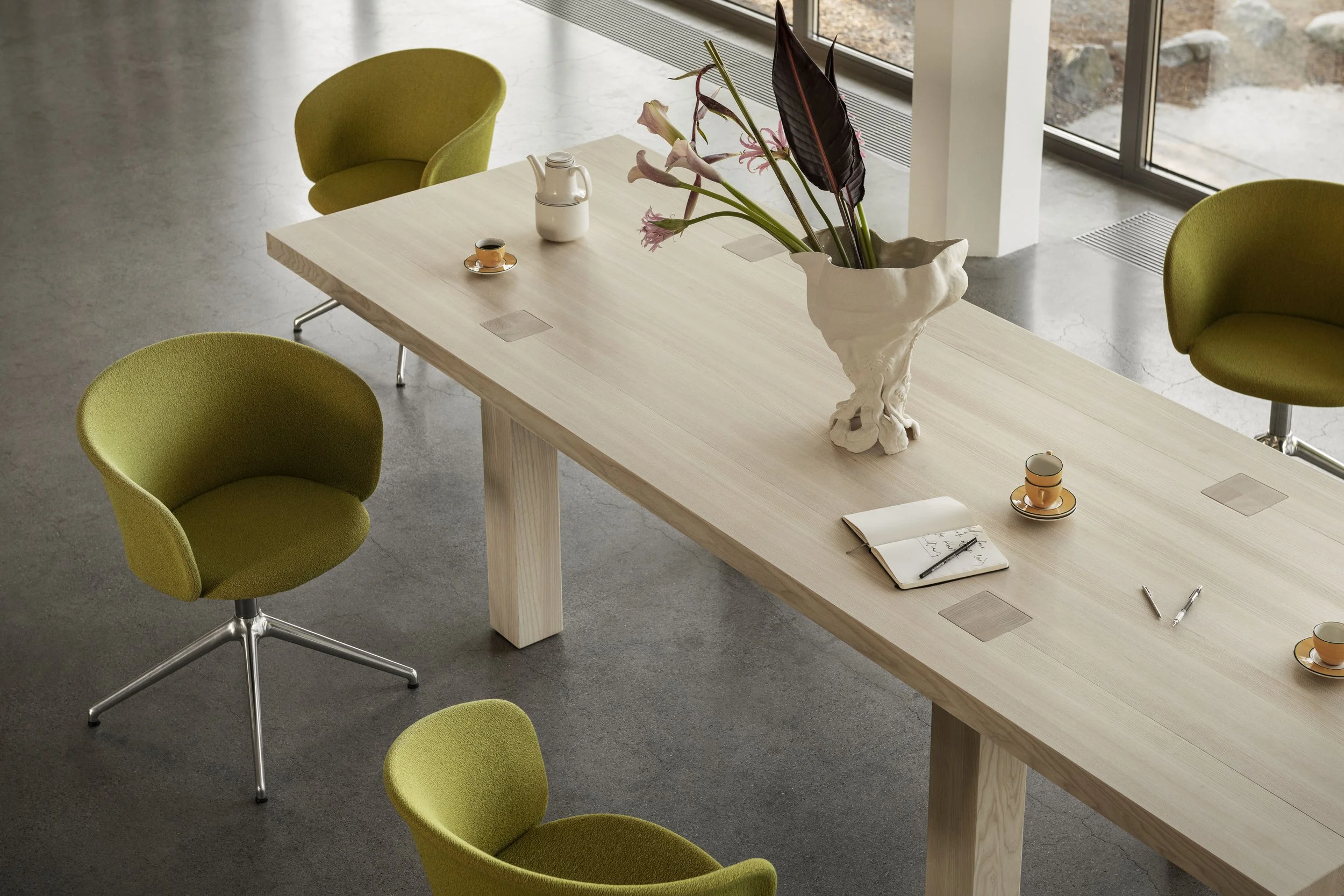 A minimalist office or conference room with a long wooden table, four green upholstered chairs, a ceramic vase with large pink and dark purple leaves, a white teapot, cups, and food items, with large windows showing an outdoor area.