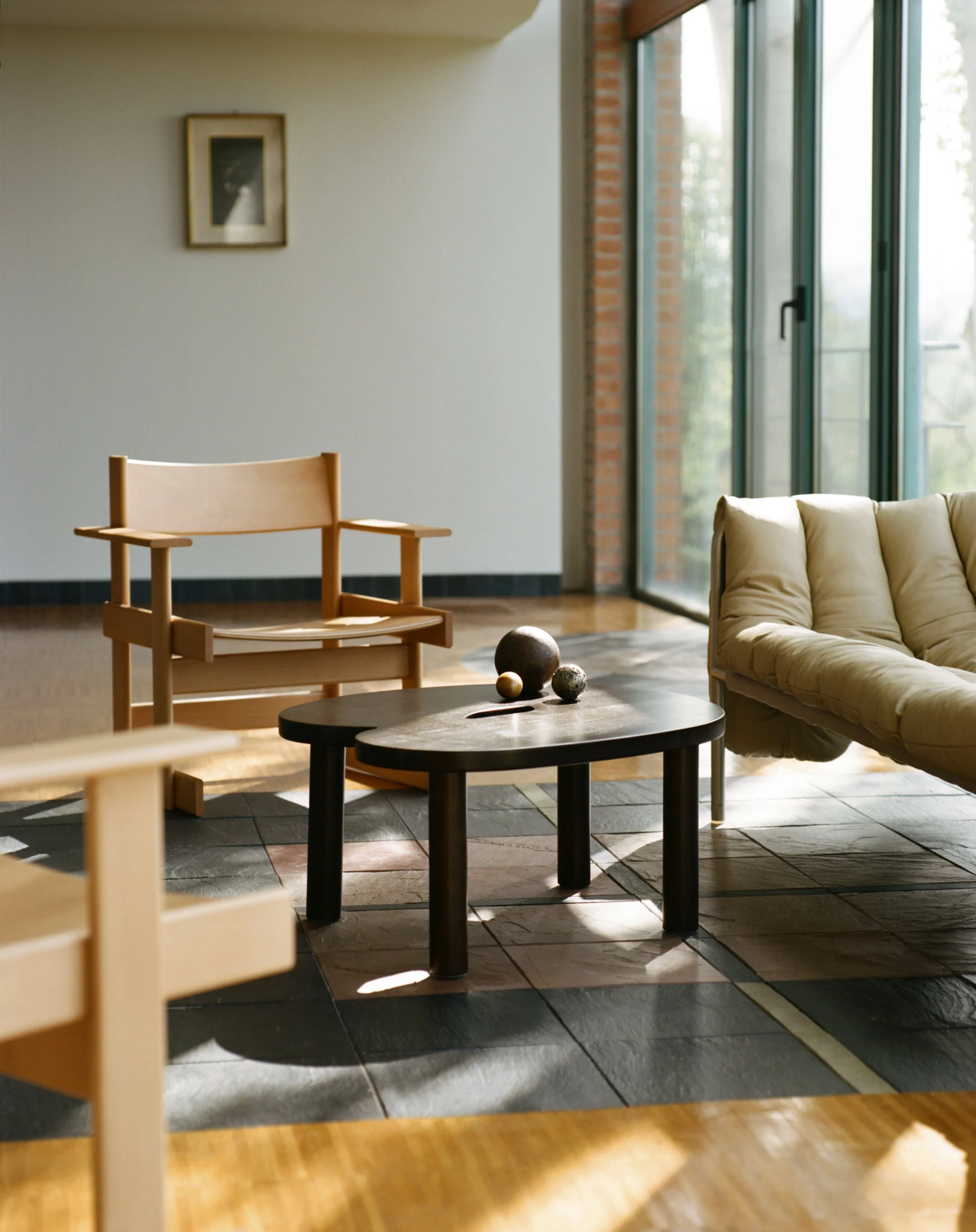 Interior view of a modern living room with a beige sofa, a black coffee table with decorative spheres, a wooden chair, and large windows letting in natural light.