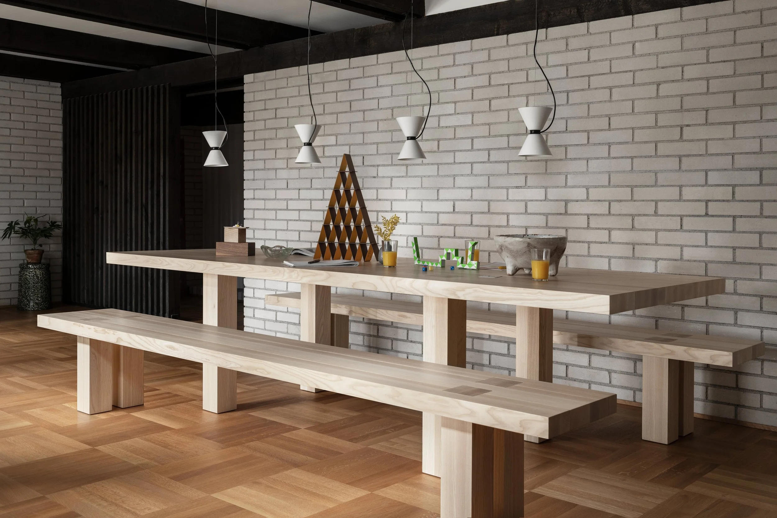 A modern interior space with a long wooden table and bench, a white brick wall, pendant lights hanging overhead, and decorative items like a small potted plant and colorful blocks on the table, with wooden flooring.