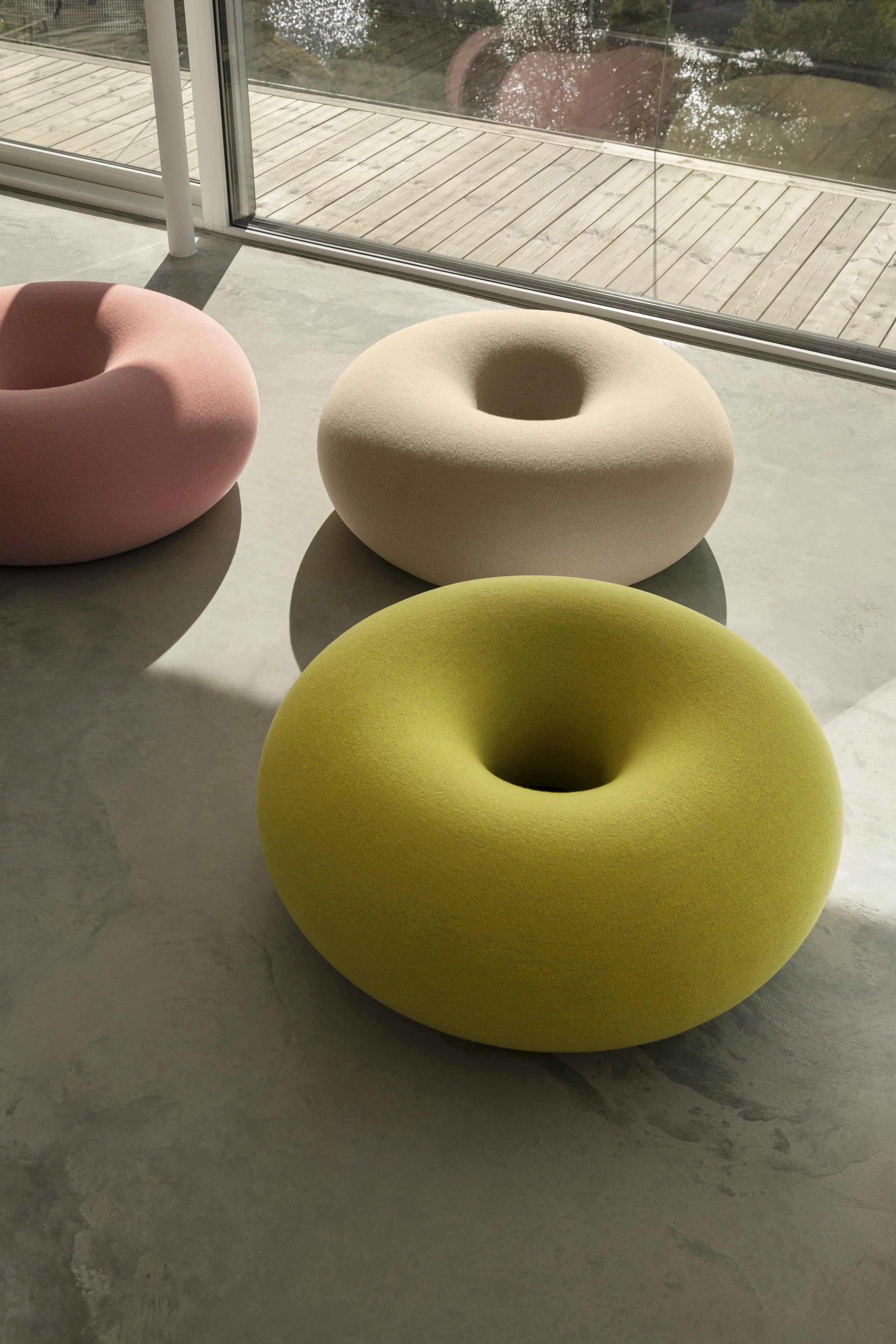 Three modern, circular, cushioned chairs in pastel colors—pink, beige, and yellow—placed on a concrete floor near a large floor-to-ceiling window with a view of a wooden deck and trees outside.