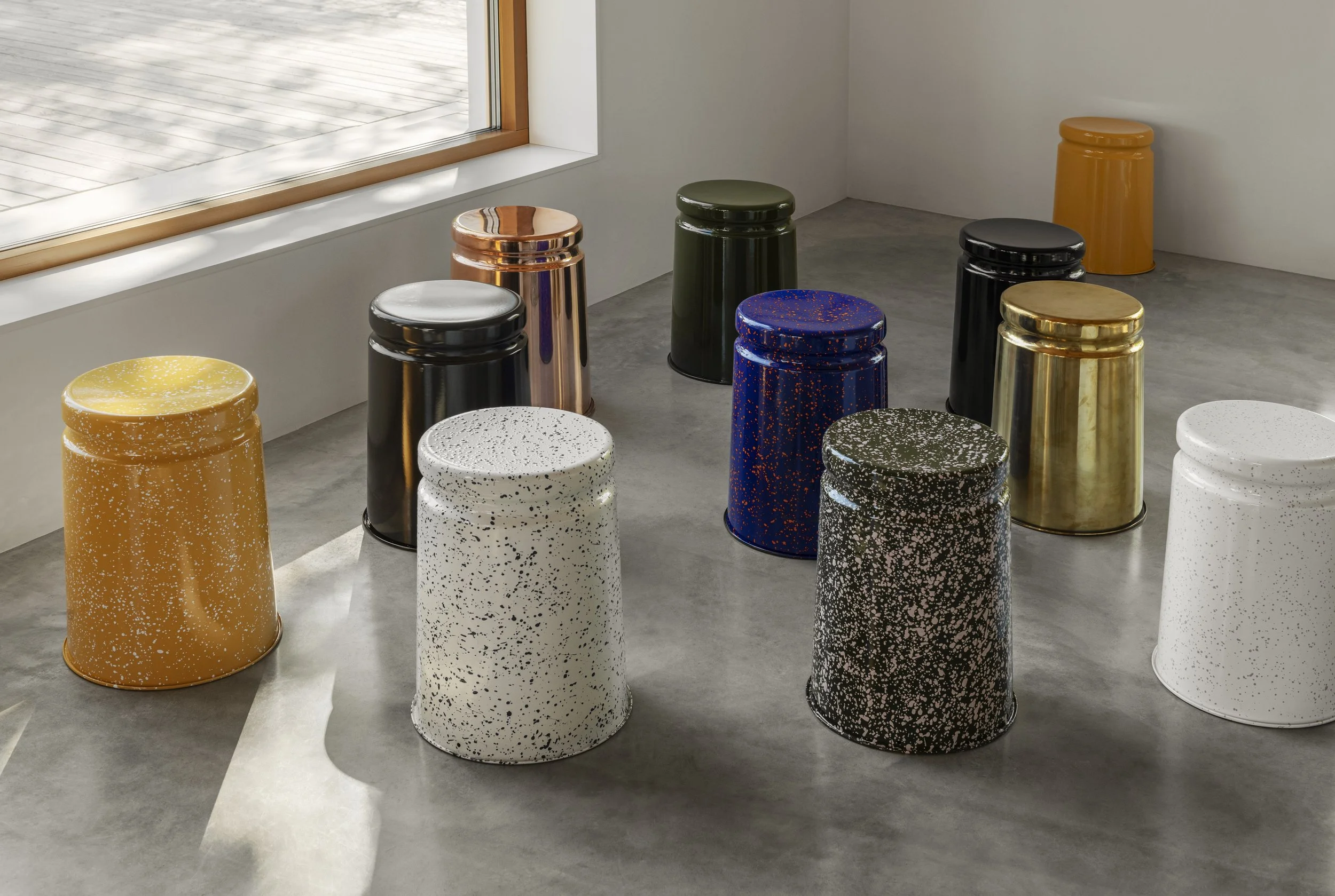 Gallery display of colorful, speckled ceramic stools in yellow, white, black, green, gold, copper, and blue, arranged near a window with wooden trim.