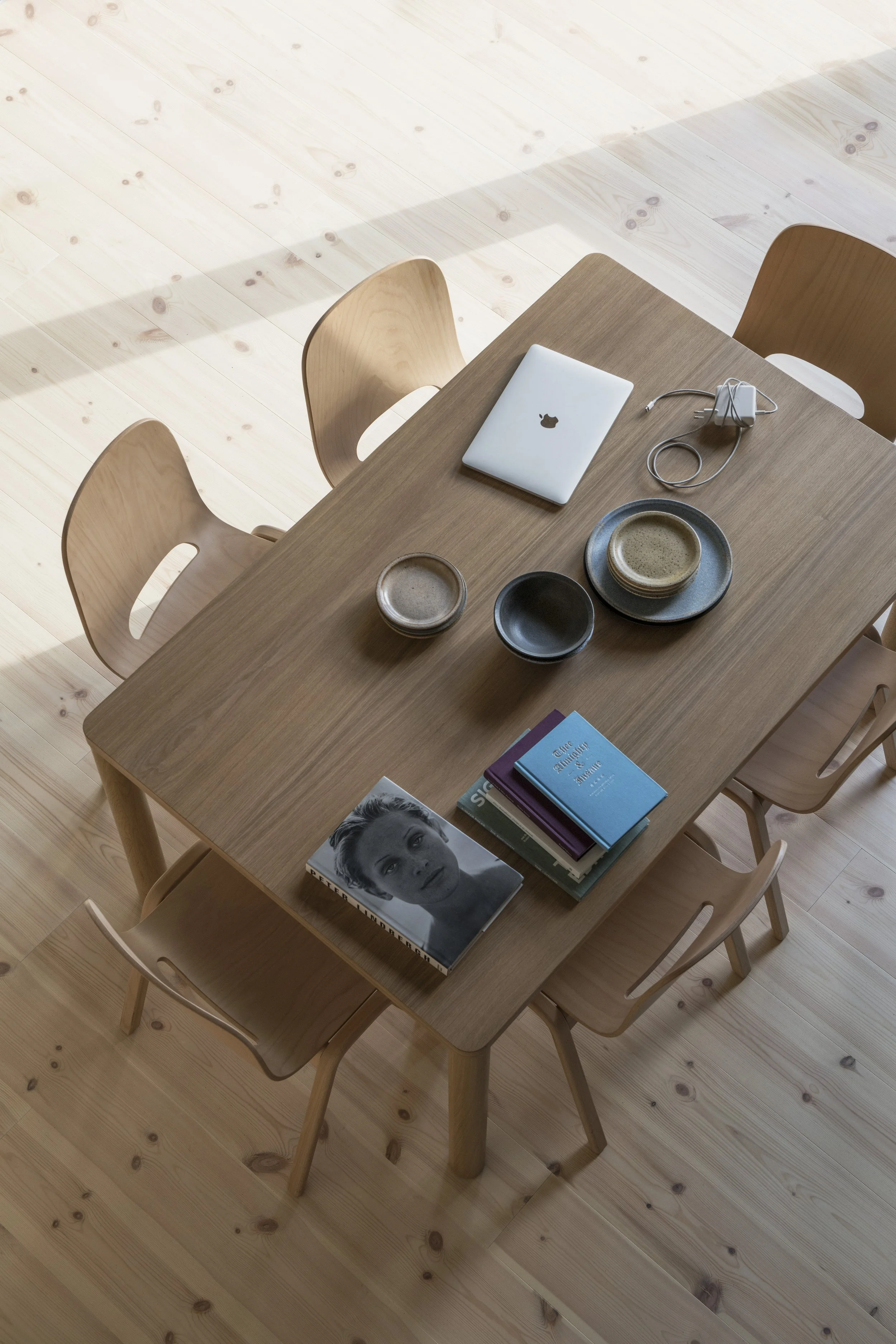 A wooden table with three chairs, a closed MacBook, a charger, three bowls, and a stack of books, including a black and white portrait of a woman on top.