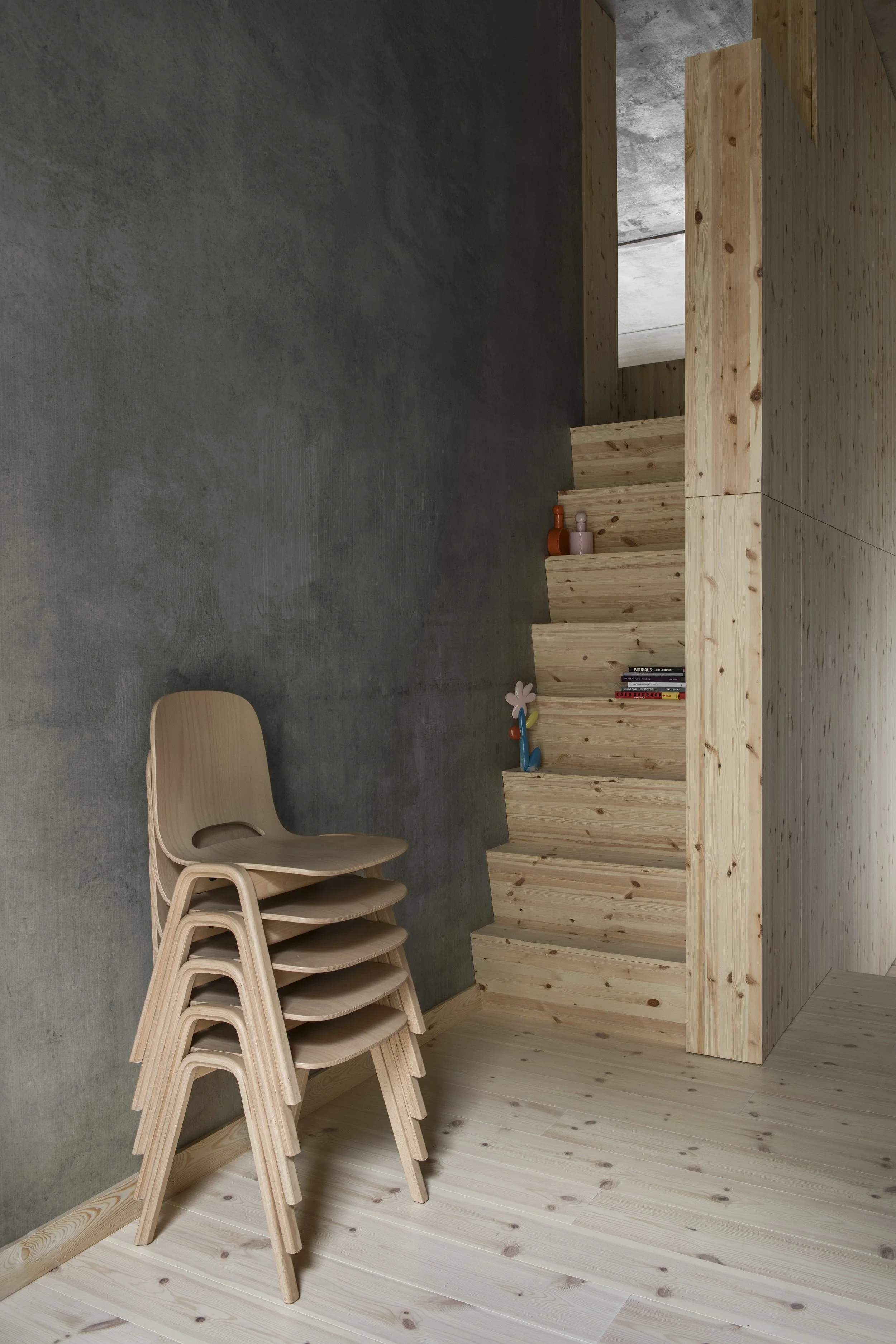 Stacked wooden chairs leaning against a gray wall, with a wooden shelving unit containing books, vases, and a small decorative item, in a modern minimalist room.
