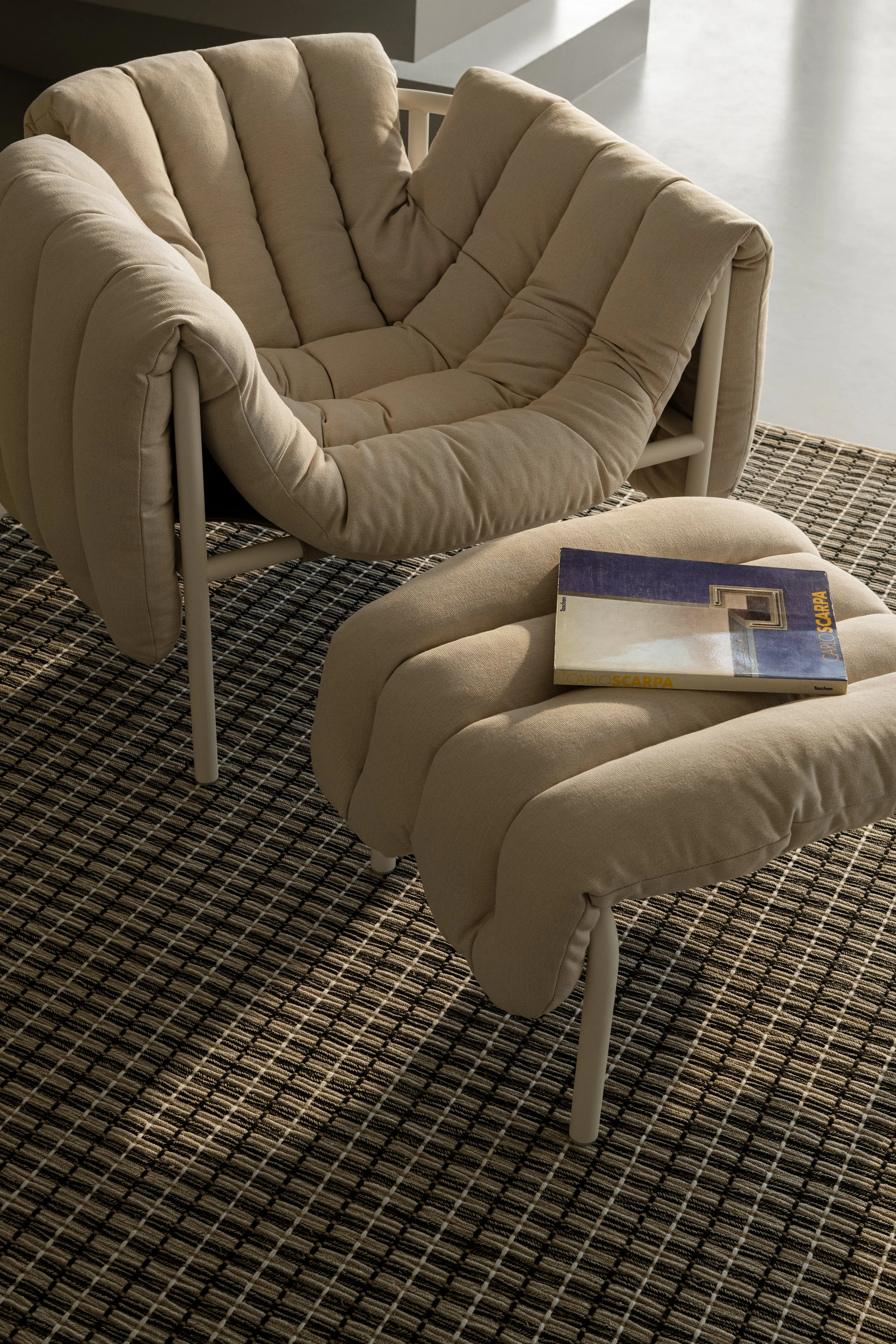 A beige cushioned armchair with a matching ottoman on a patterned black and brown rug in a modern interior space.
