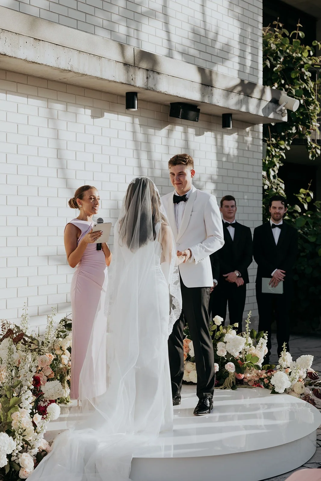 A wedding ceremony taking place outdoors with a bride and groom exchanging vows, surrounded by floral arrangements, with an officiant and three groomsmen in the background.