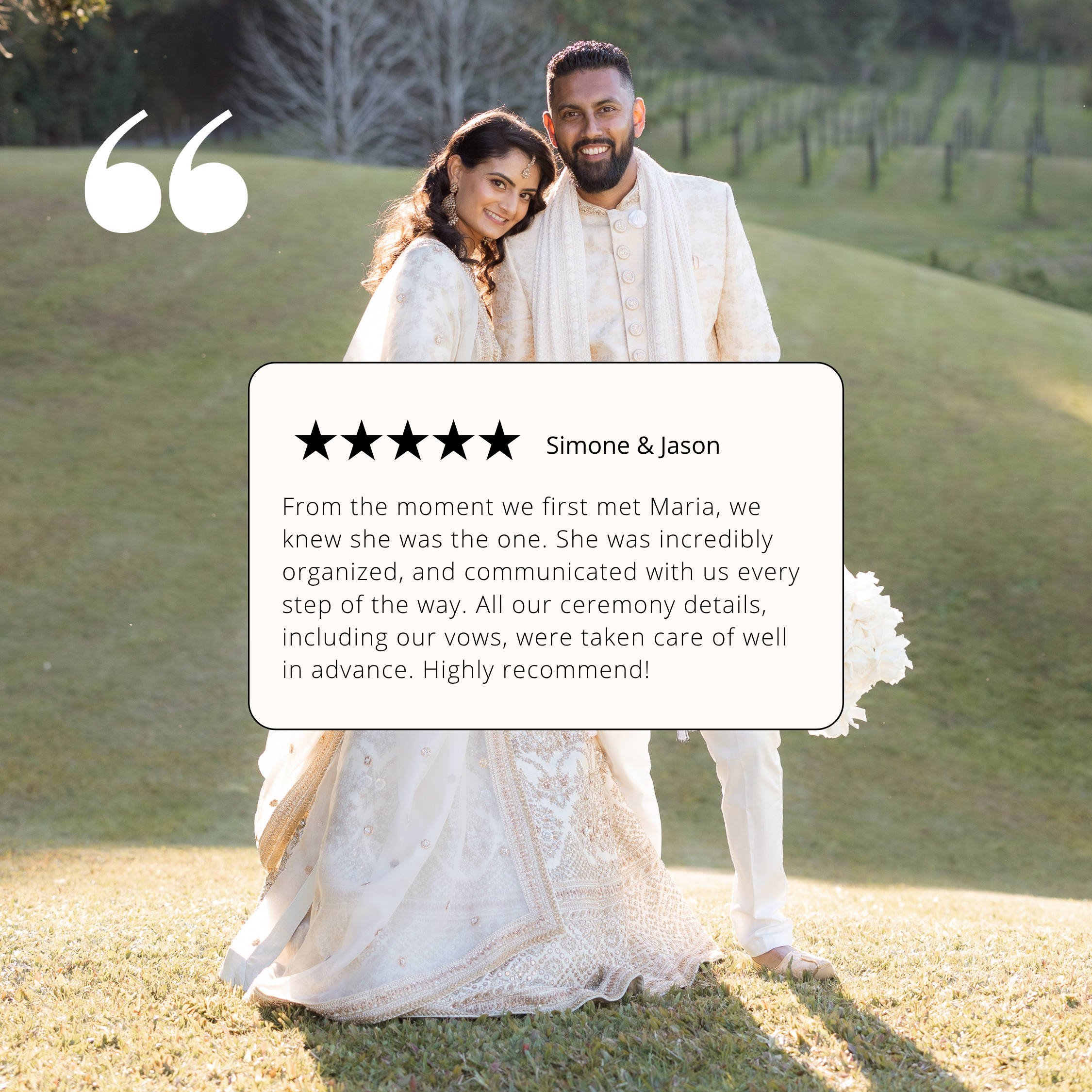A smiling bride and groom in traditional cream-colored wedding attire outdoors during daytime, standing on grass with a background of trees and a gentle hillside.