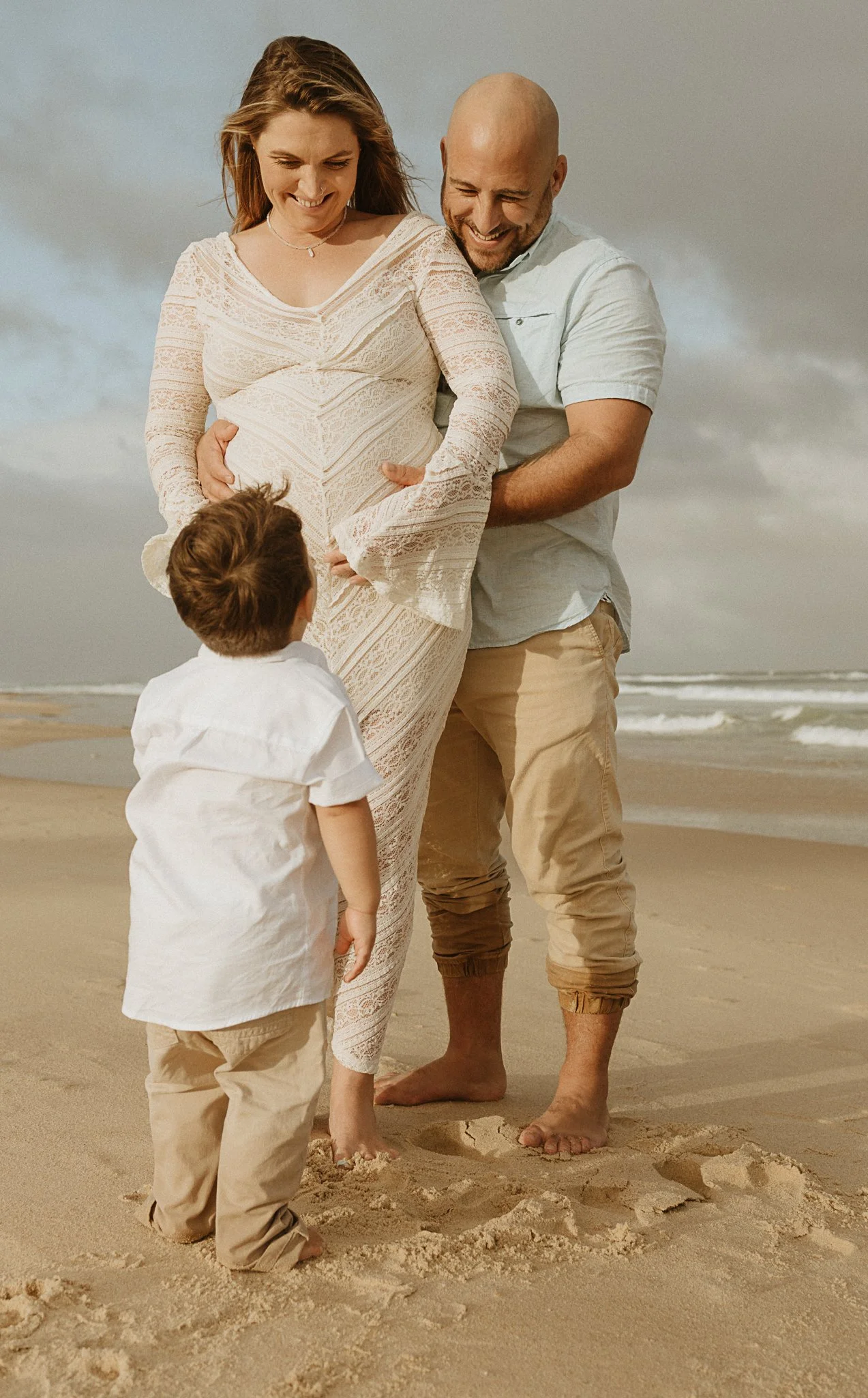 Smiling pregnant woman in a lace dress, her partner, and a young child on the beach during daytime, all smiling and enjoying the moment of Maternity session on Caves Beach Lake Macquarie/