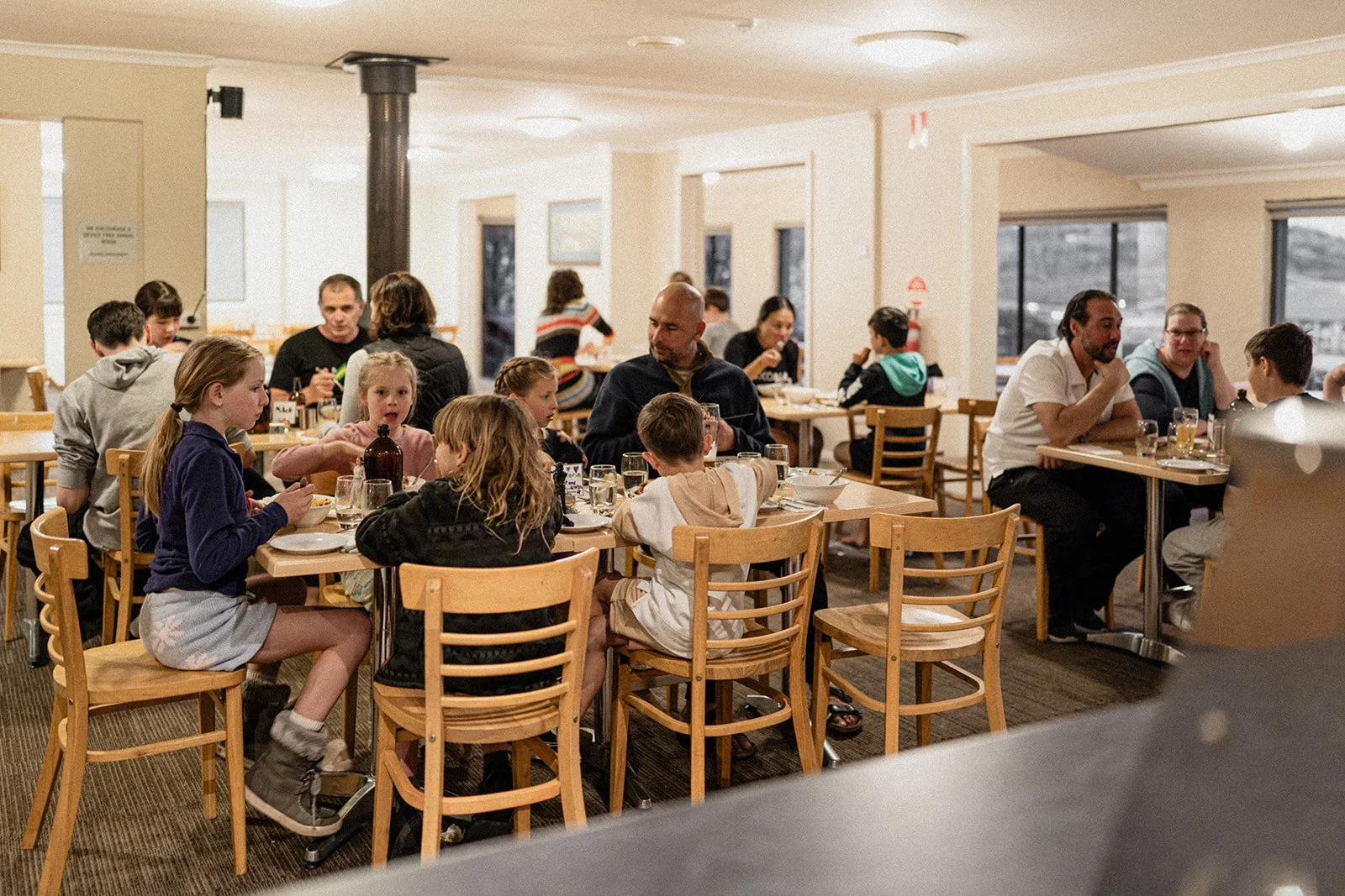 People dining at tables in a restaurant, including children and adults, with large windows and bright lighting.