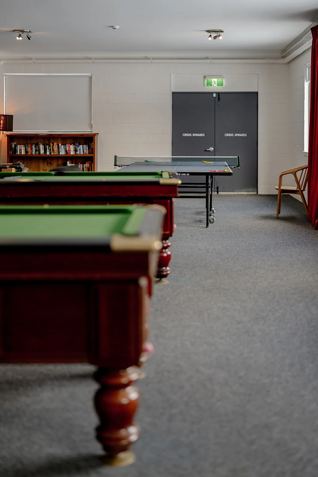 View of a recreational room with pool table in the foreground, ping pong table in the background, bookshelves on the left, a wooden chair on the right, and a gray door with 'Opens Inwards' sign and an emergency exit sign above it.