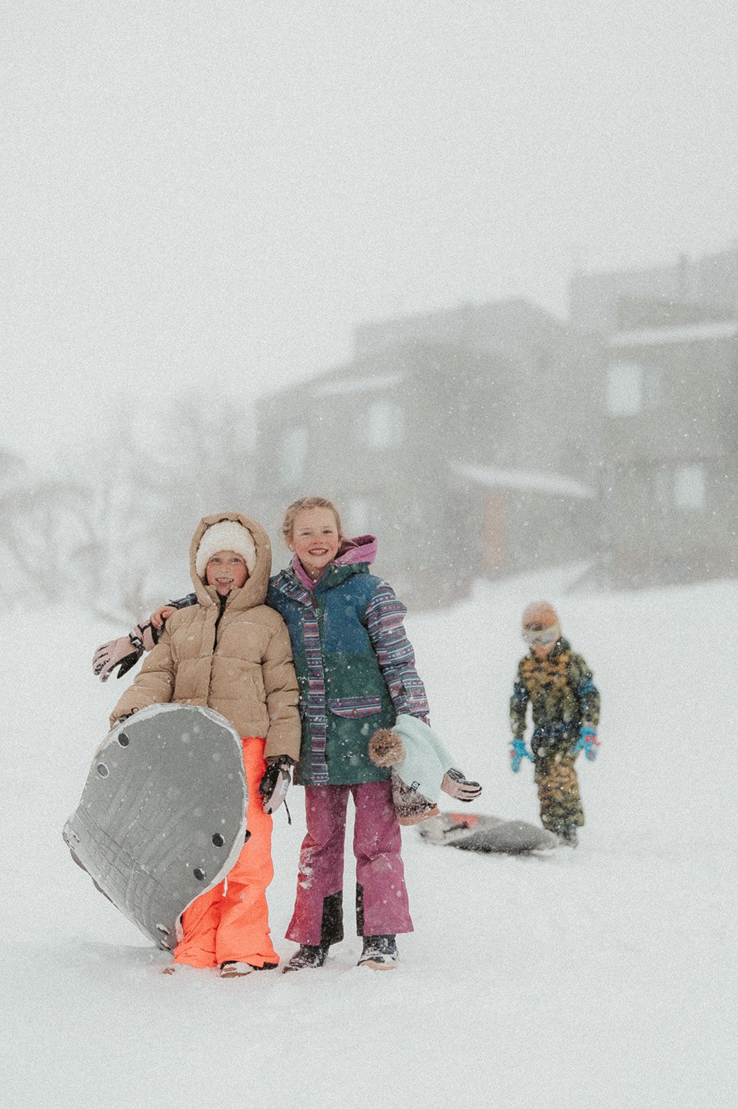 Two children in winter clothing smiling and posing with a sled in a snowy landscape, with another child in the background playing in the snow and a house or building in the distance.