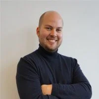 Lächelnder Mann mit Bart und schwarzen Rollkragenpullover vor weißem Hintergrund.