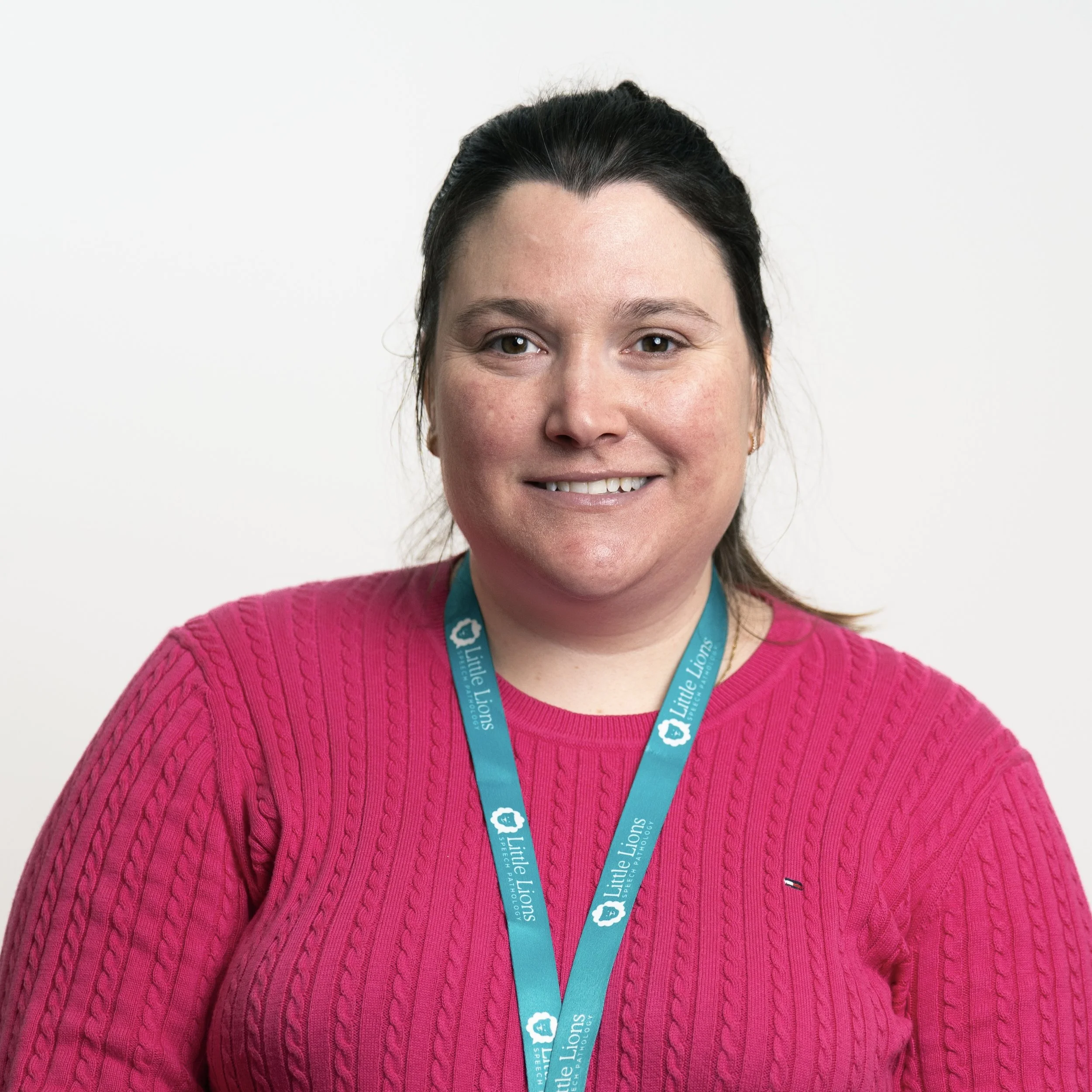 A woman wearing a pink sweater and a blue lanyard with the 'Little Lions' logo, smiling at the camera against a plain white background.