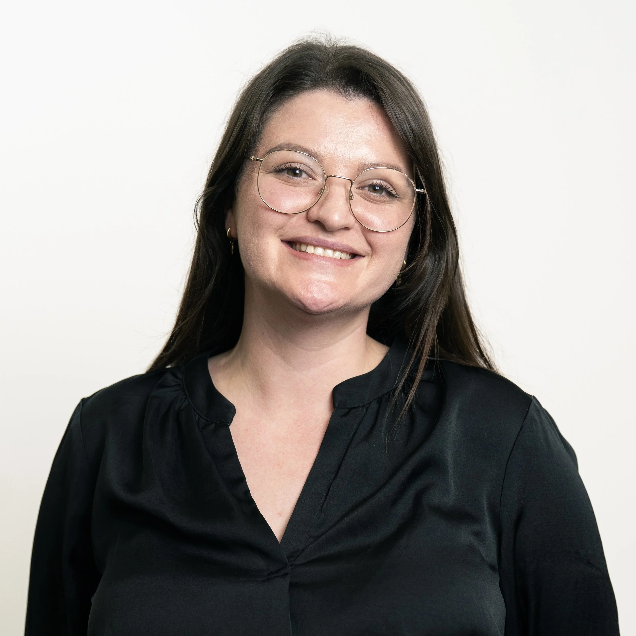 A smiling woman with long dark hair, glasses, and a black shirt standing against a plain white background.